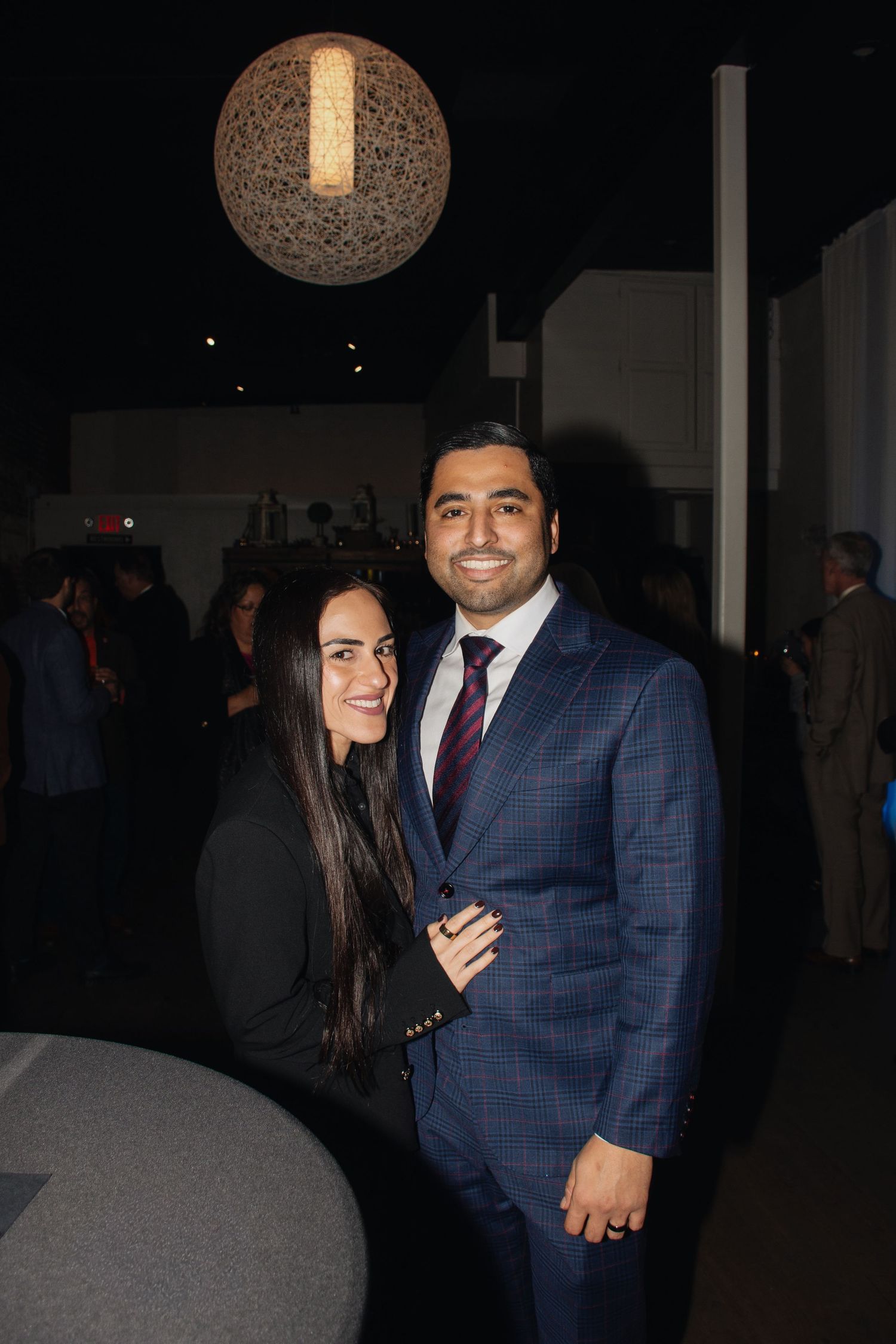 A couple smiles together at an evening event, with the person in a navy checkered suit and another in dark attire.