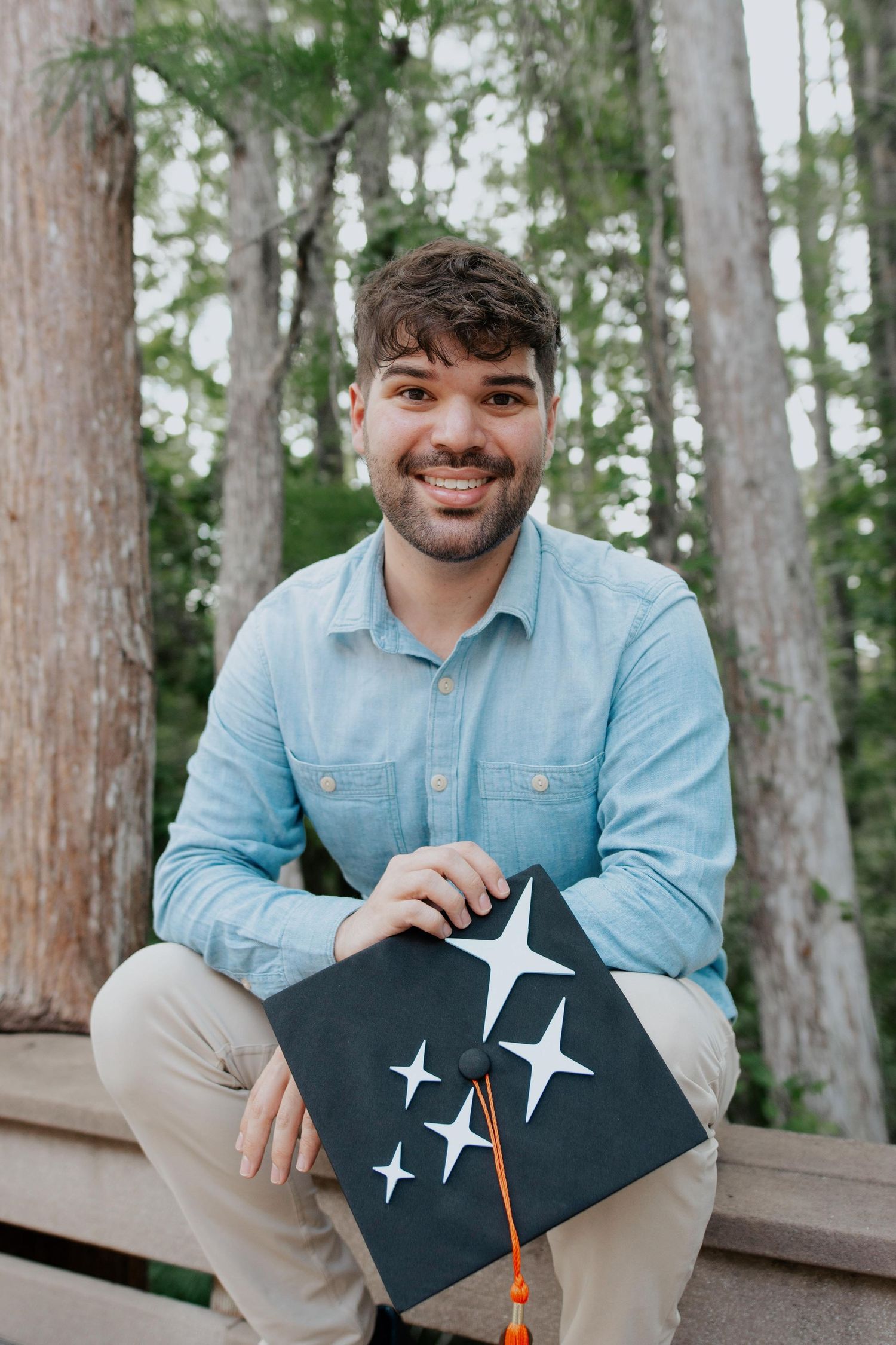 A person in a light blue denim shirt holds a graduation cap with decorative stars on top in a wooded outdoor setting.
