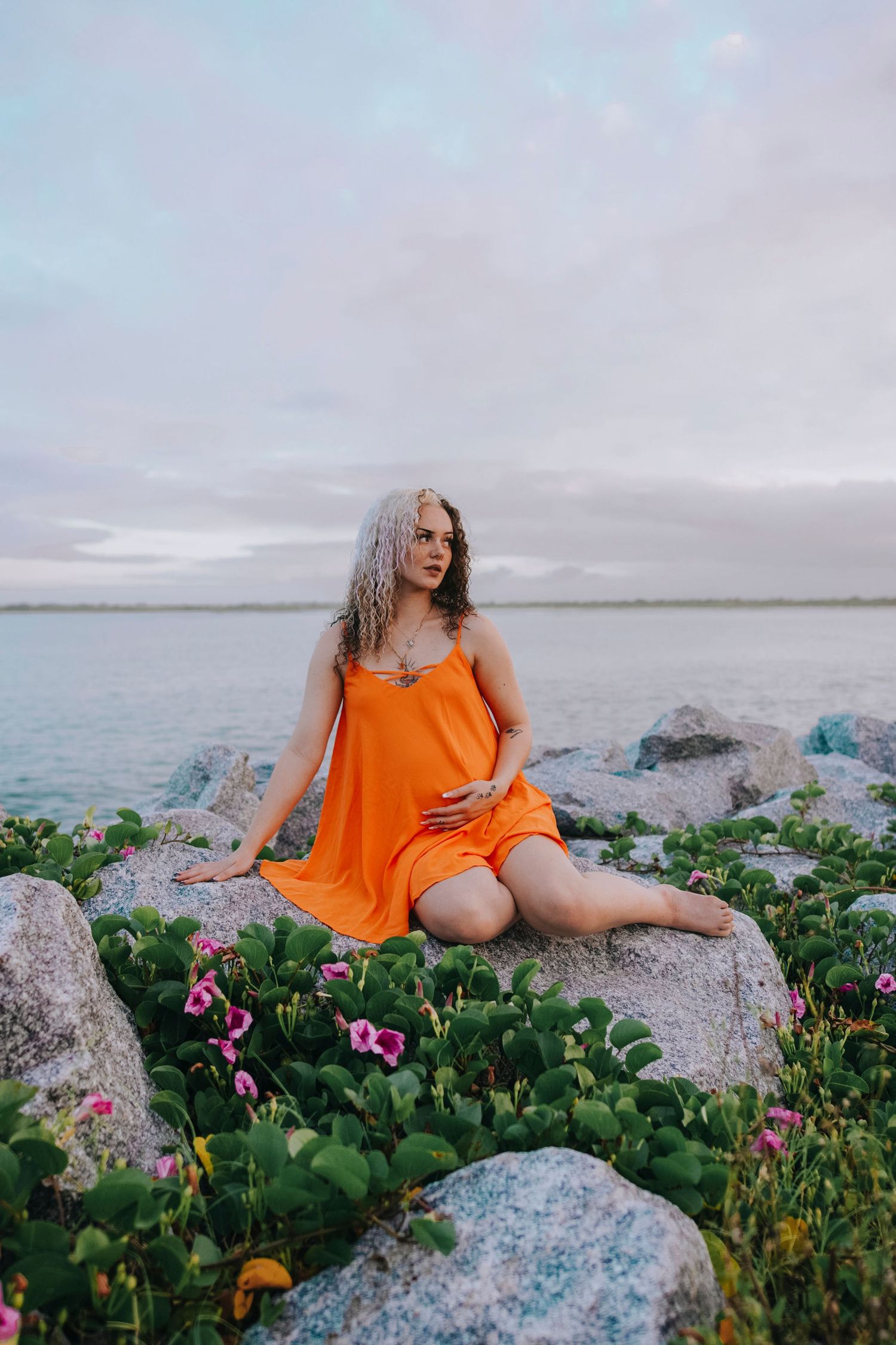 Woman in bright orange dress poses on coastal rocks surrounded by wildflowers.