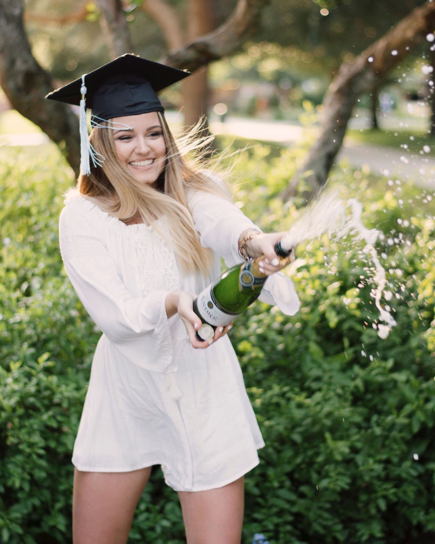 Graduate in cap and gown pops champagne during outdoor celebration photoshoot.