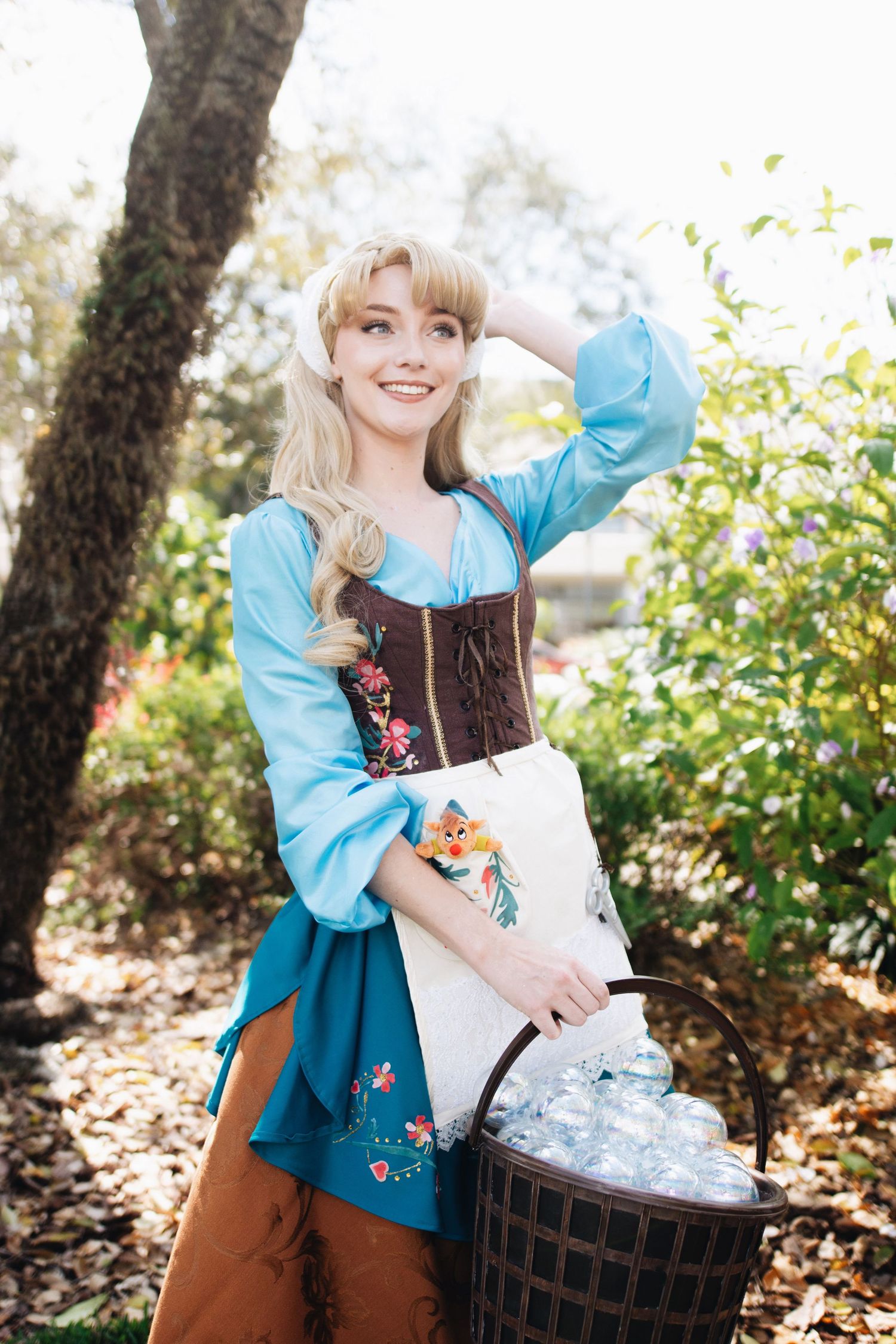 A cosplayer in a peasant-style costume with turquoise dress and white apron carries a basket outdoors.