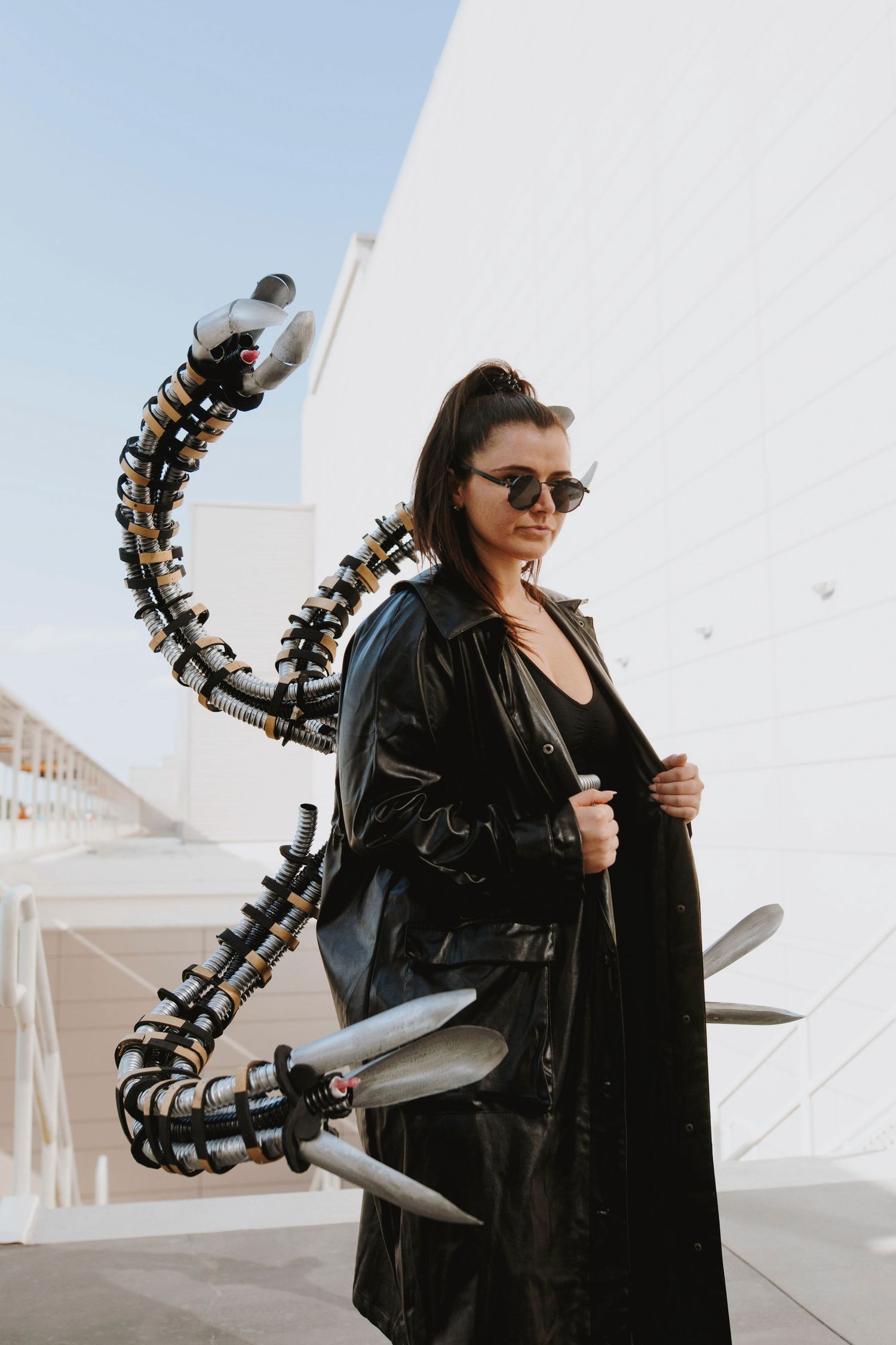 Dramatic black leather outfit with mechanical tentacle prop against white architecture.