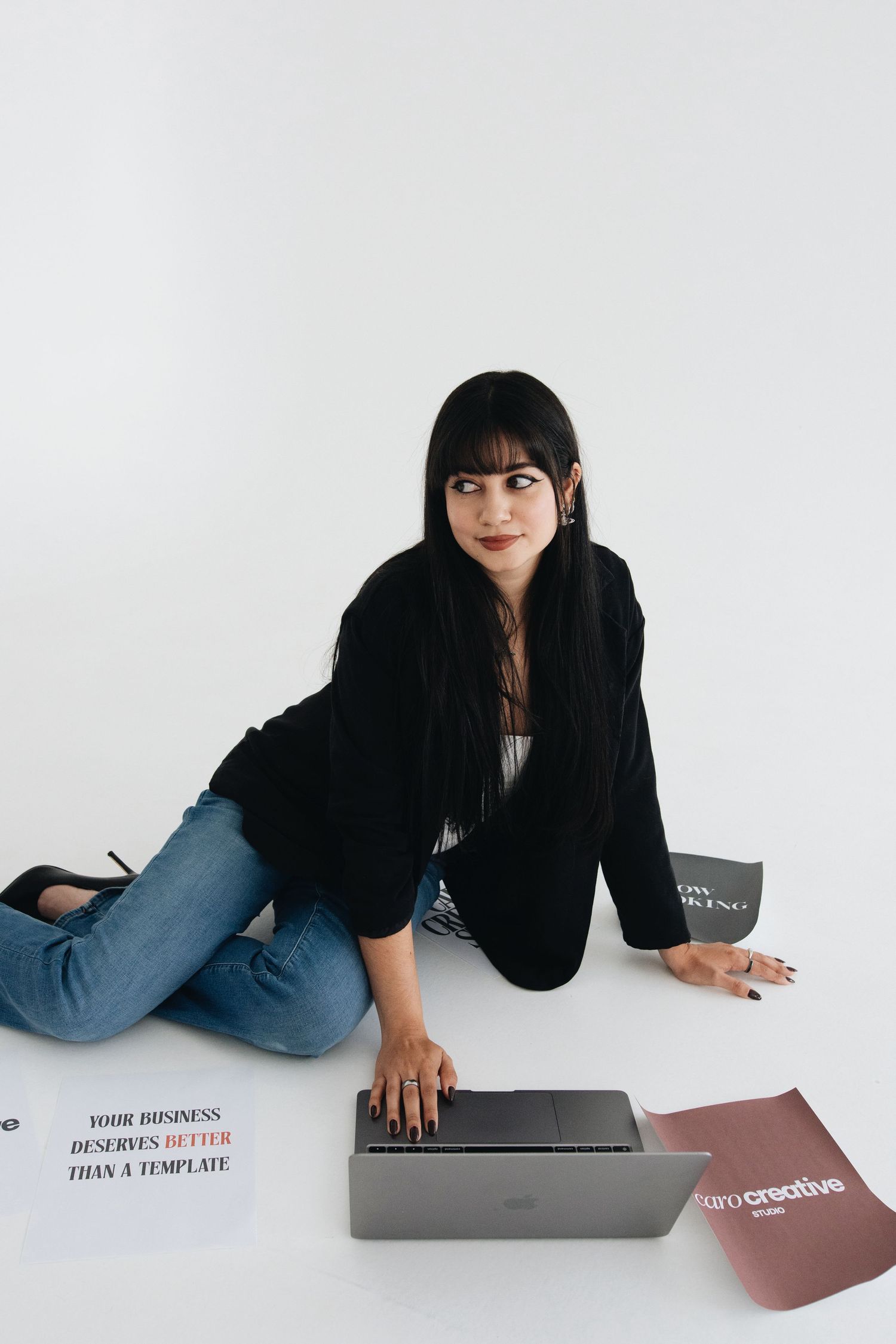 Woman in black blazer and jeans sits on floor with laptop, surrounded by business papers on white background.