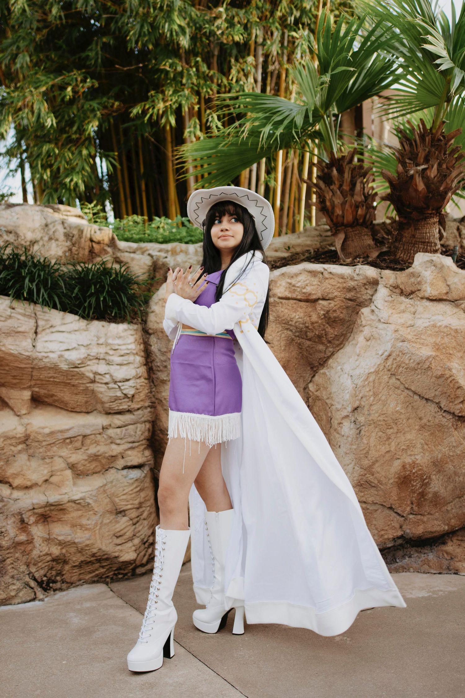 Cosplay photoshoot featuring white boots and purple outfit against tropical rock garden setting.