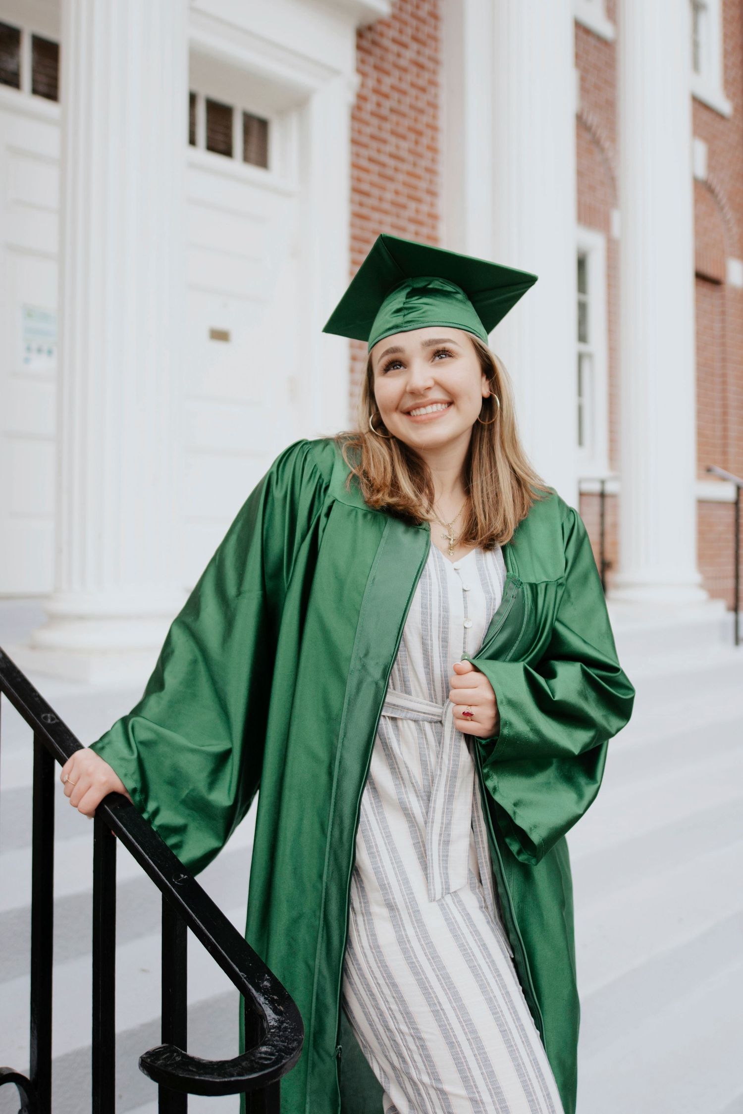 Graduate in green cap and gown poses on brick building steps with white columns.