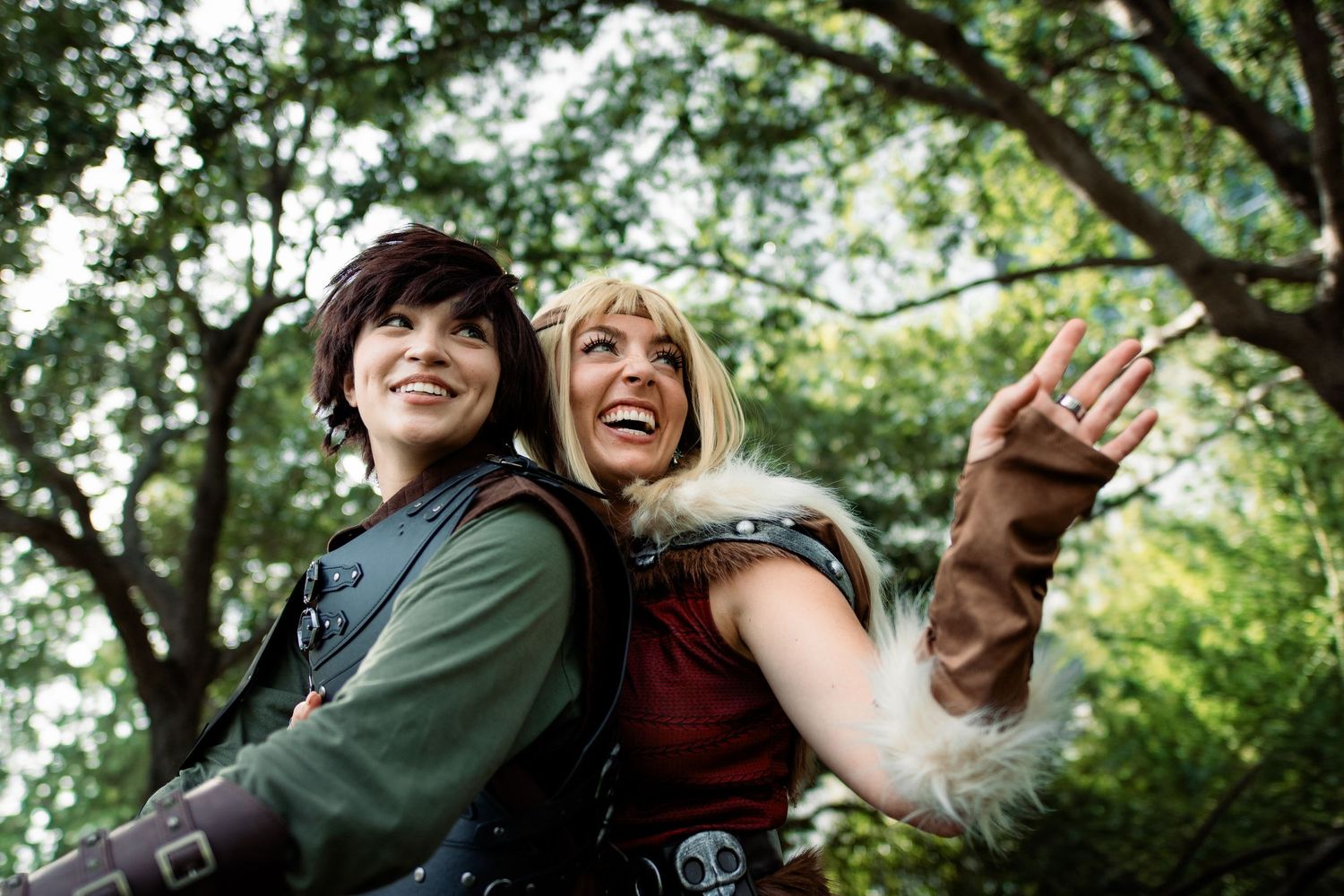 Two cosplayers in medieval fantasy costumes pose outdoors under green trees, smiling and pointing upward.