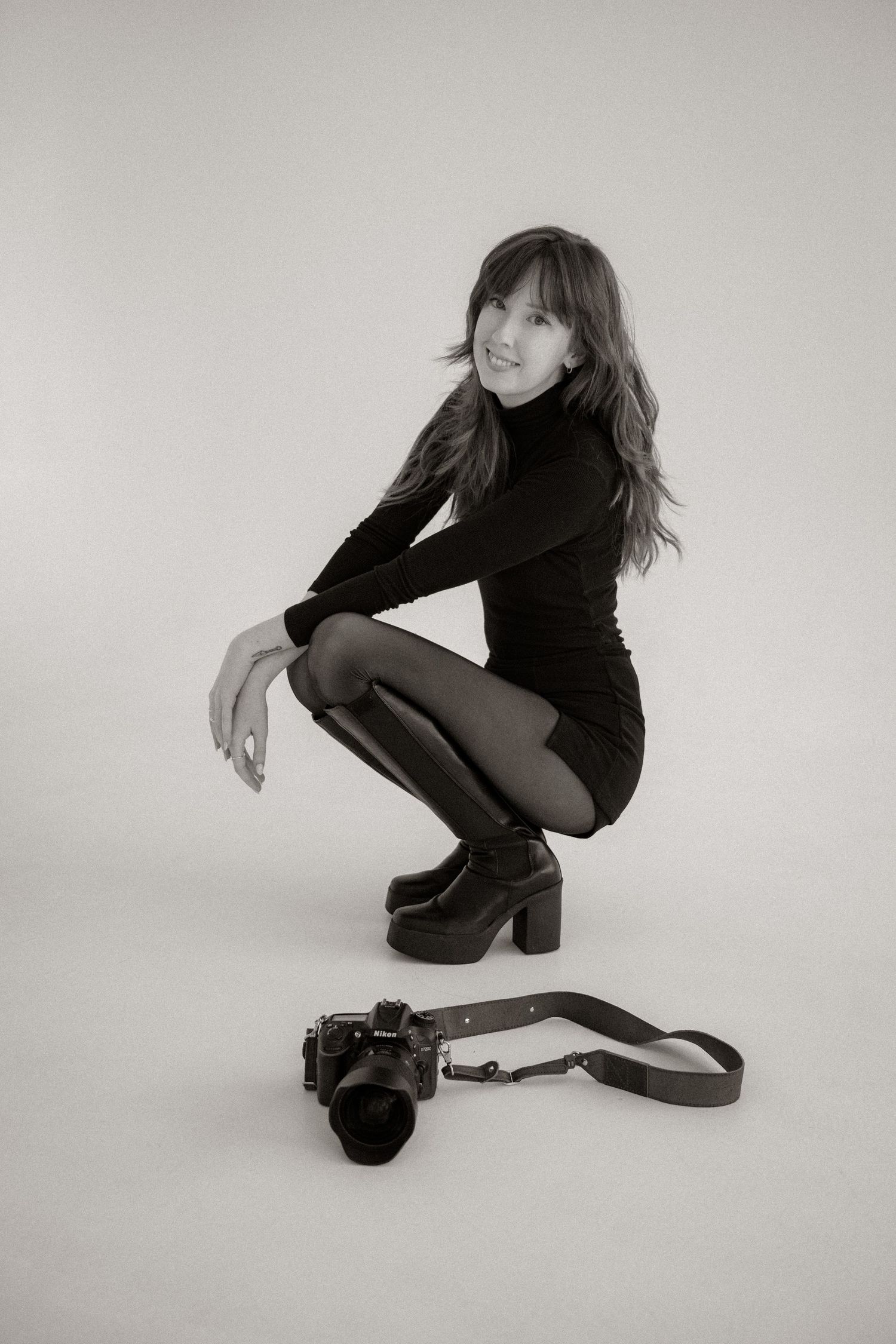 Black and white photo of a young woman crouching in a black outfit with platform boots beside a camera.