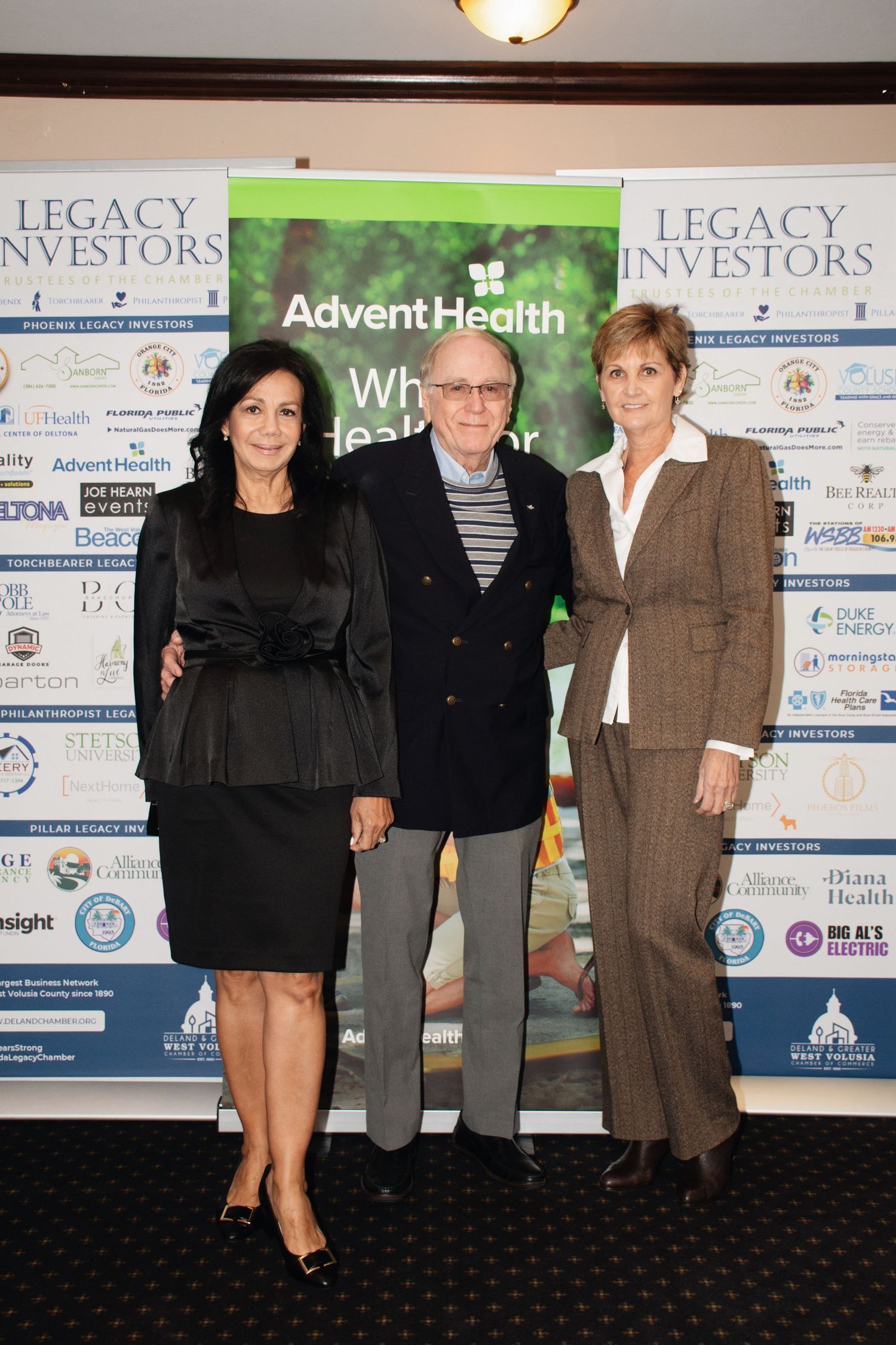 A group of people standing in front of corporate banners displaying Legacy and AdventHealth logos at a business event.