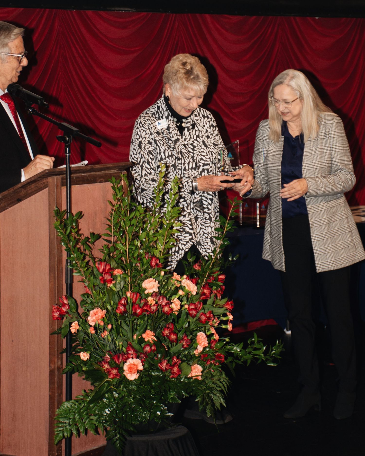 Two people stand near floral arrangements with red curtains in the background during an indoor event.