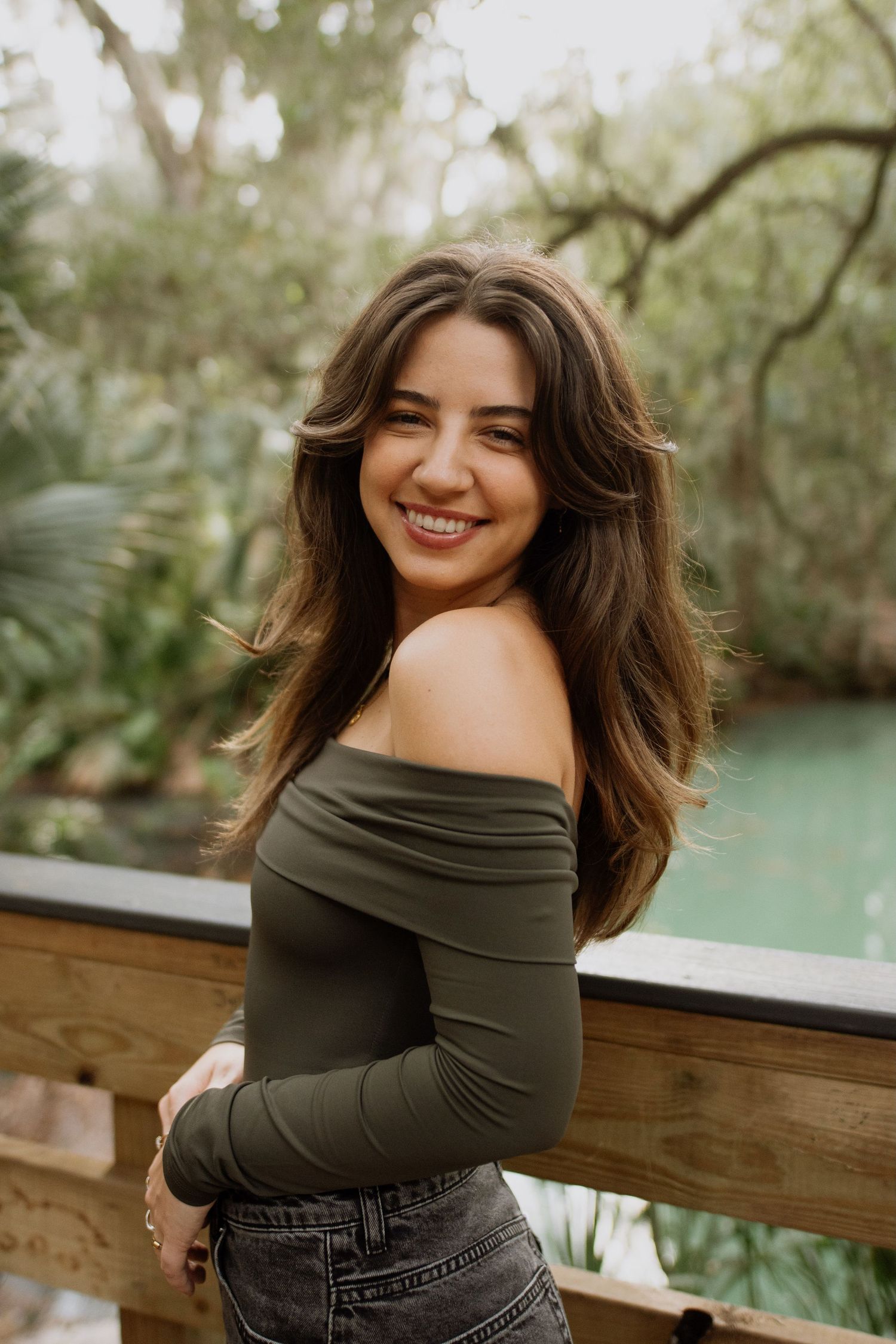 Woman in olive green off-shoulder top and jeans smiles while posing on wooden railing near tranquil water.