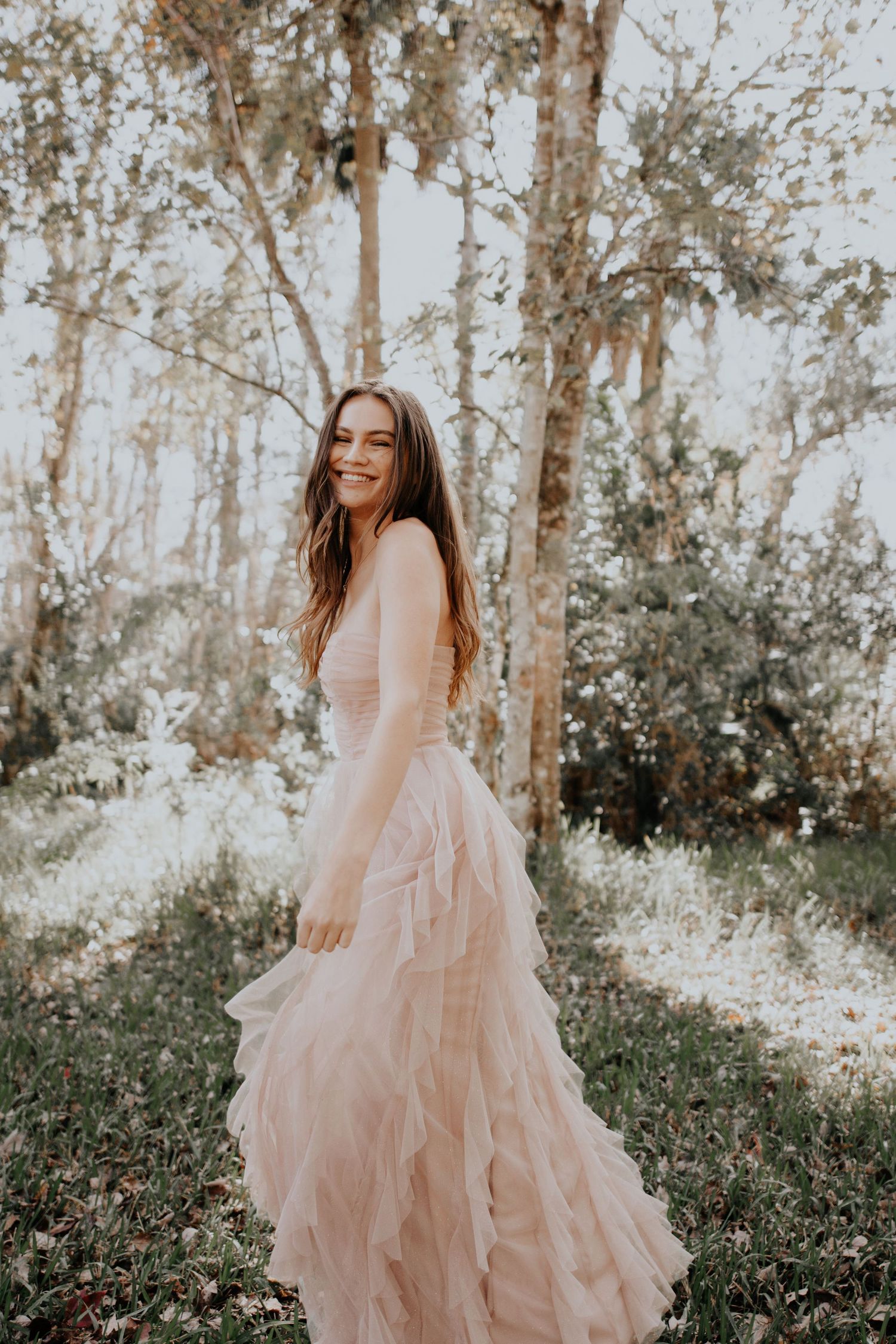 Young woman in flowing pink tulle gown twirls through sunlit forest setting.