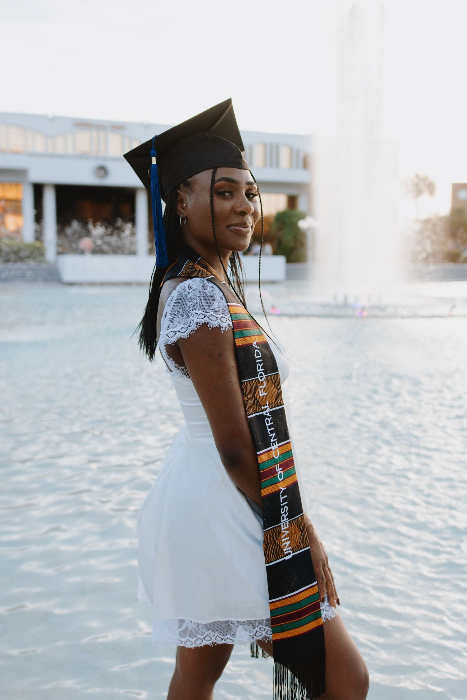Young woman in graduation cap and kente stole poses in white dress by university fountain.