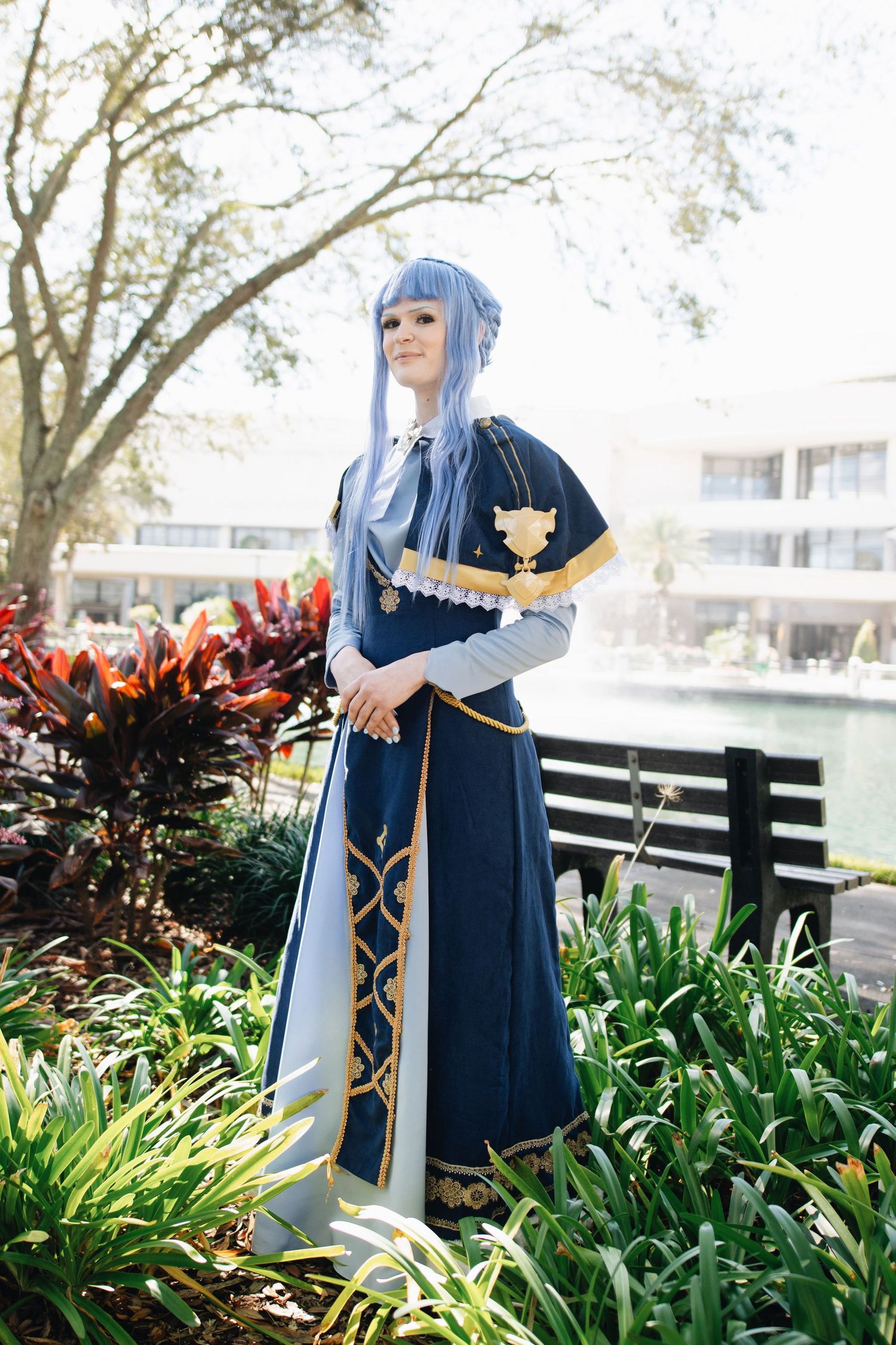 Cosplayer in dark dress with gold accents poses near flowering plants and water.