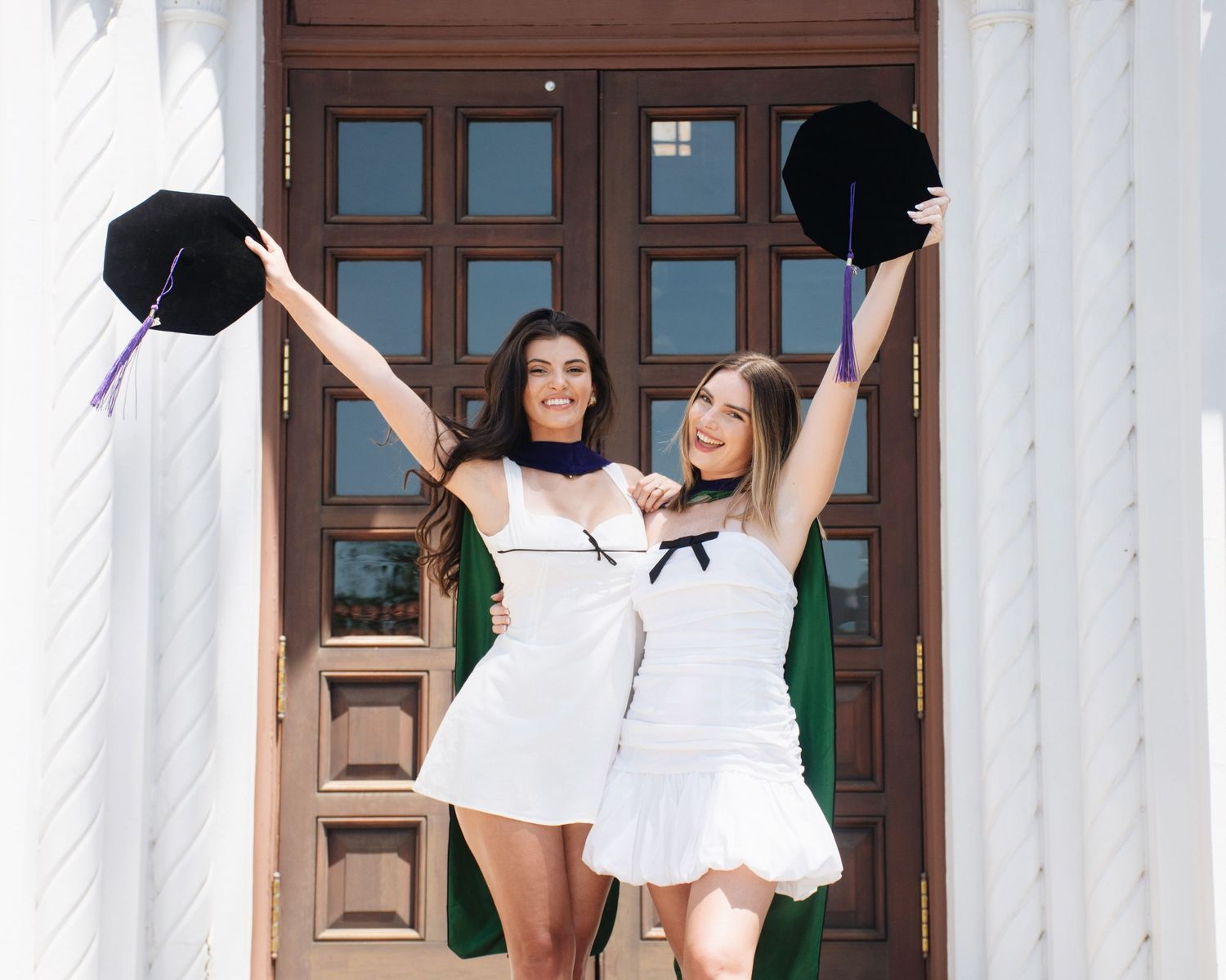 Two graduates in white dresses pose playfully in front of Stetson University College of Law entrance.