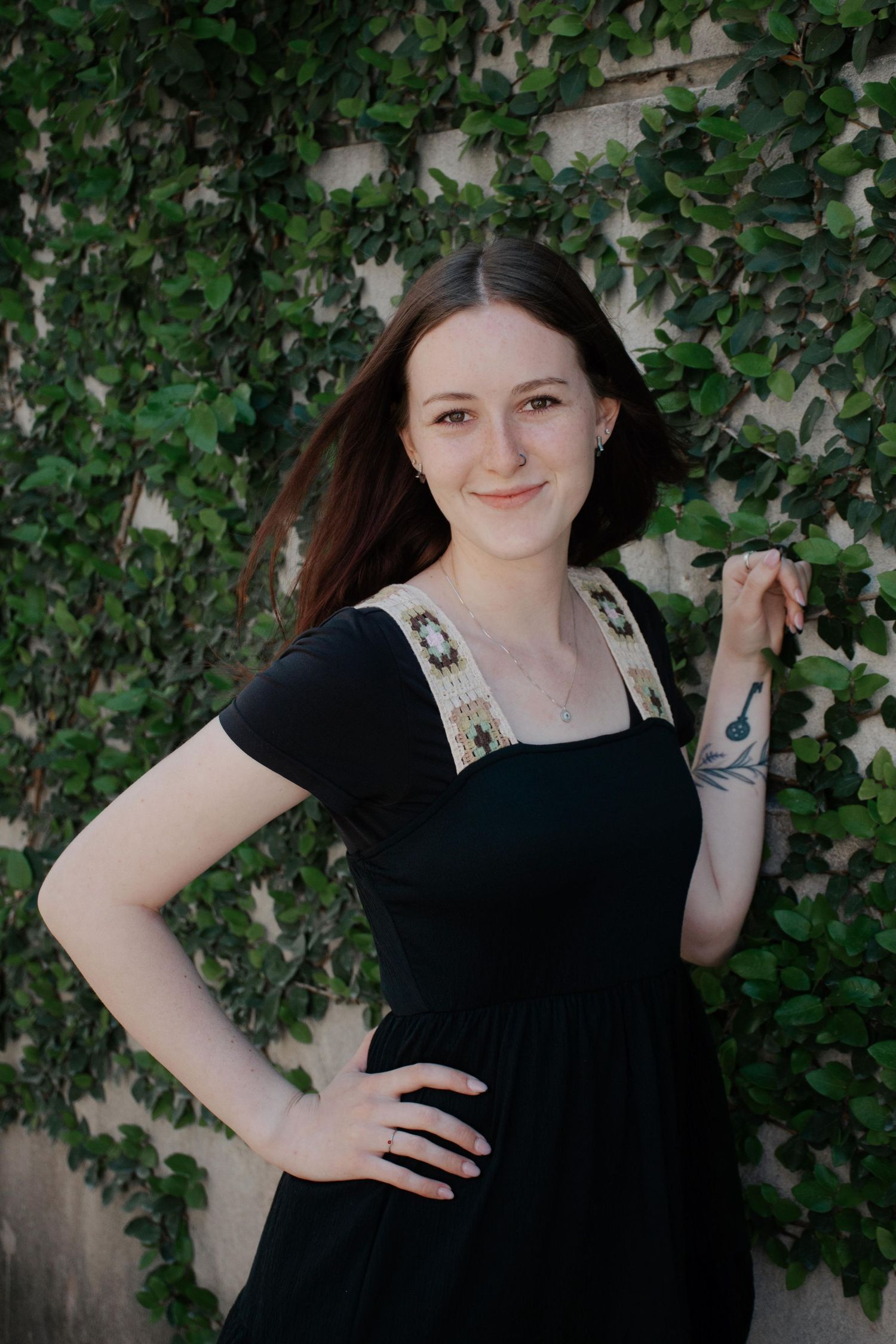 Young woman in black dress poses playfully against green vine-covered wall in garden setting.