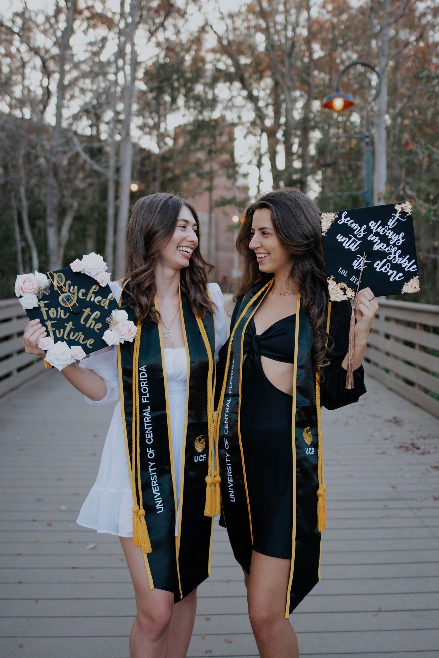 Two graduates in black caps and gowns holding decorated graduation caps on a campus walkway.