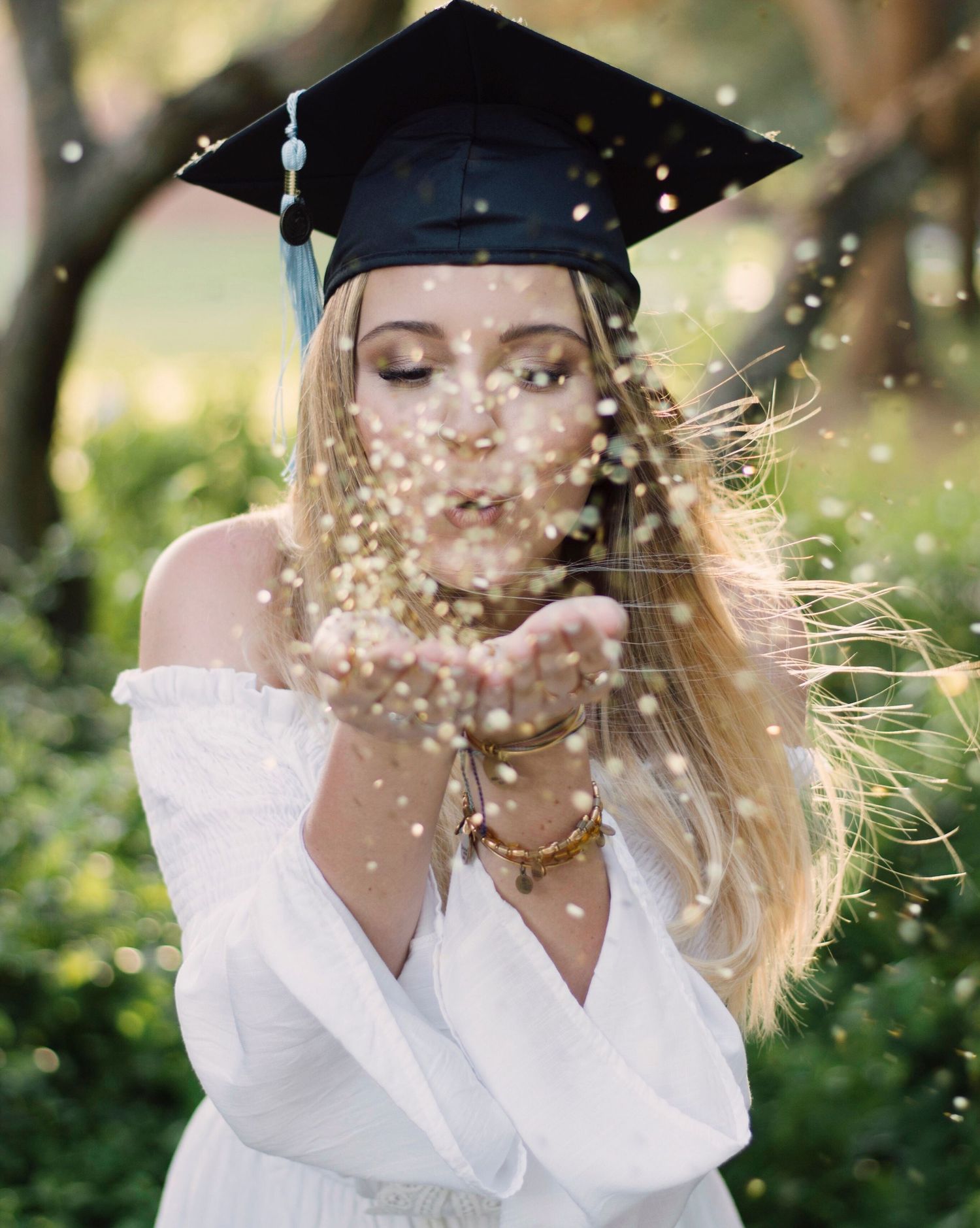Graduate in cap and gown blows glitter during outdoor graduation photoshoot.