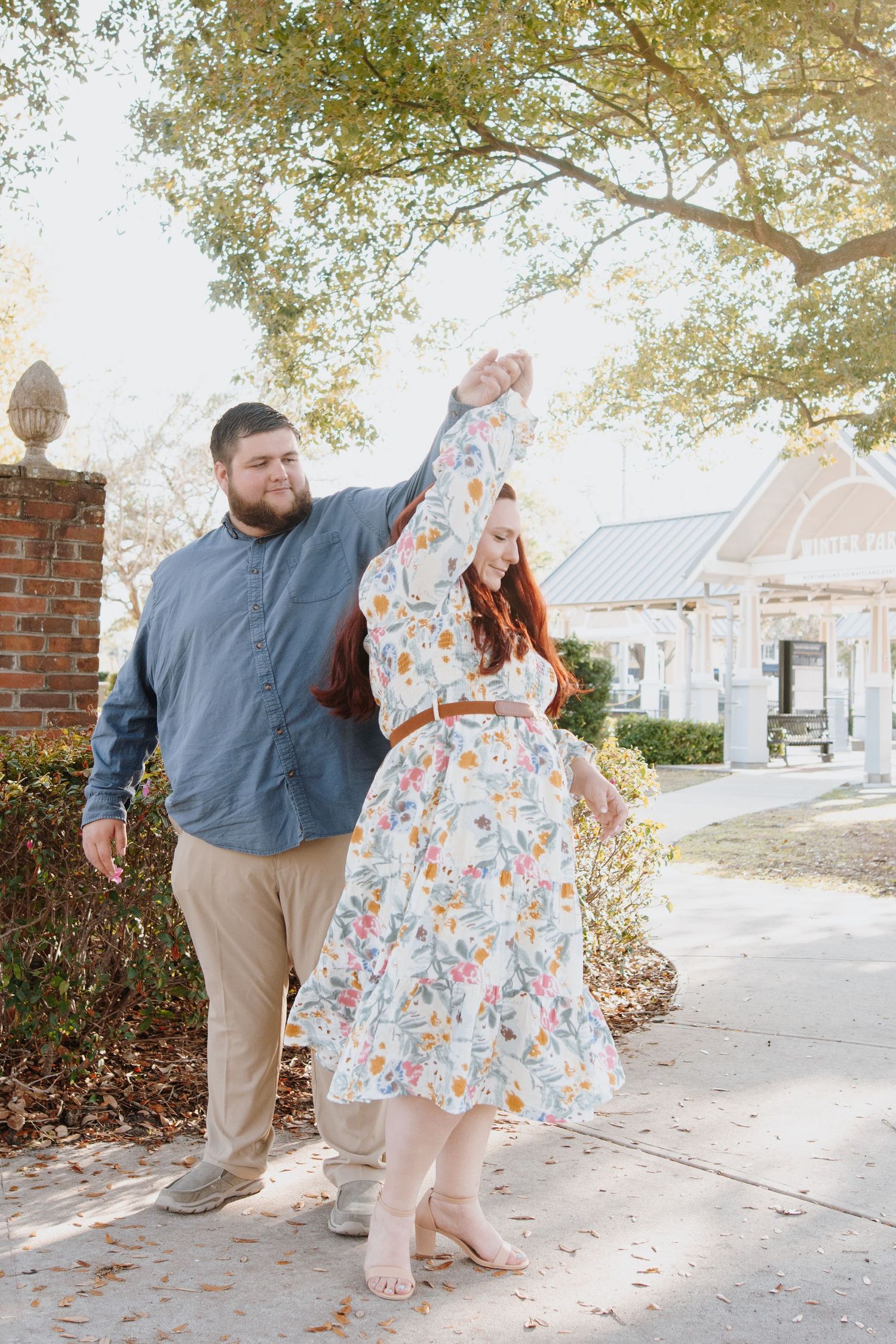 A couple in casual attire dances together on a leaf-covered sidewalk during an autumn engagement photo session.