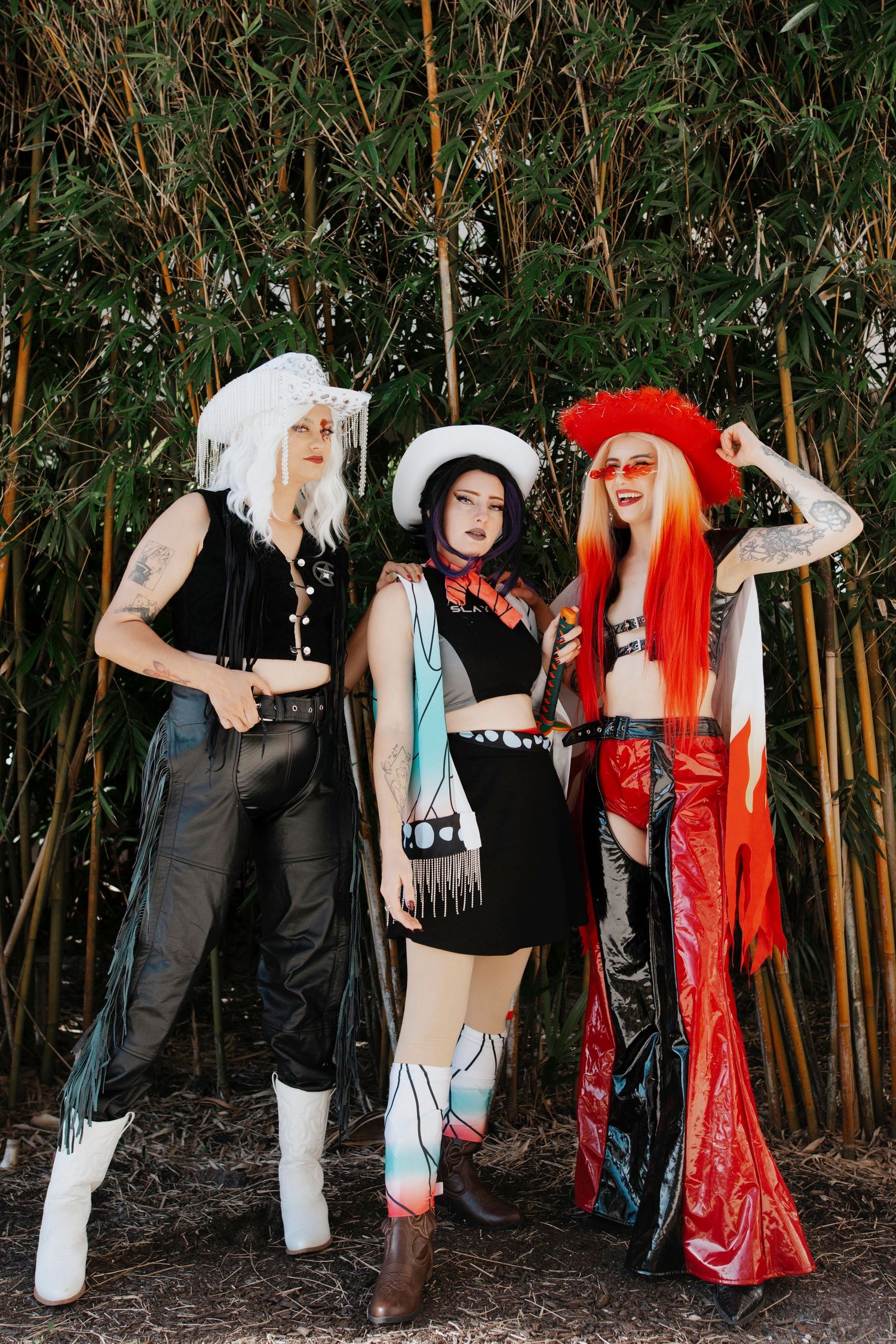 Three cosplayers pose together in front of bamboo, wearing stylized outfits with white, black and red color themes.