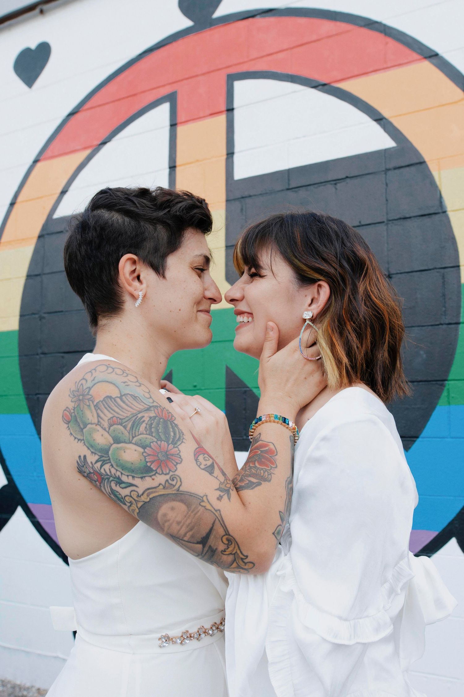 Happy couple in white outfits sharing an intimate moment against colorful rainbow mural.