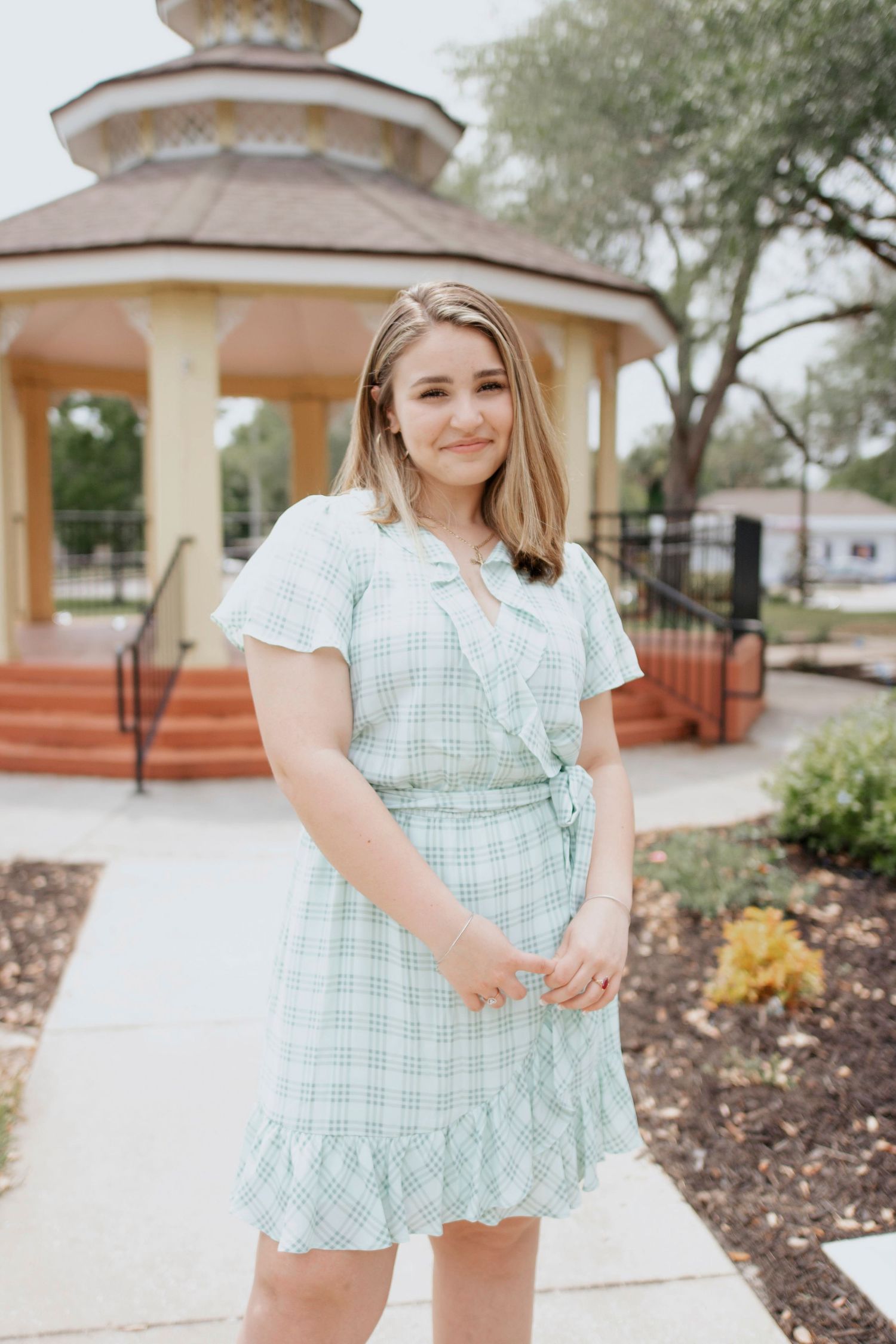 A person wearing a mint green gingham dress poses outdoors near a gazebo and landscaping.