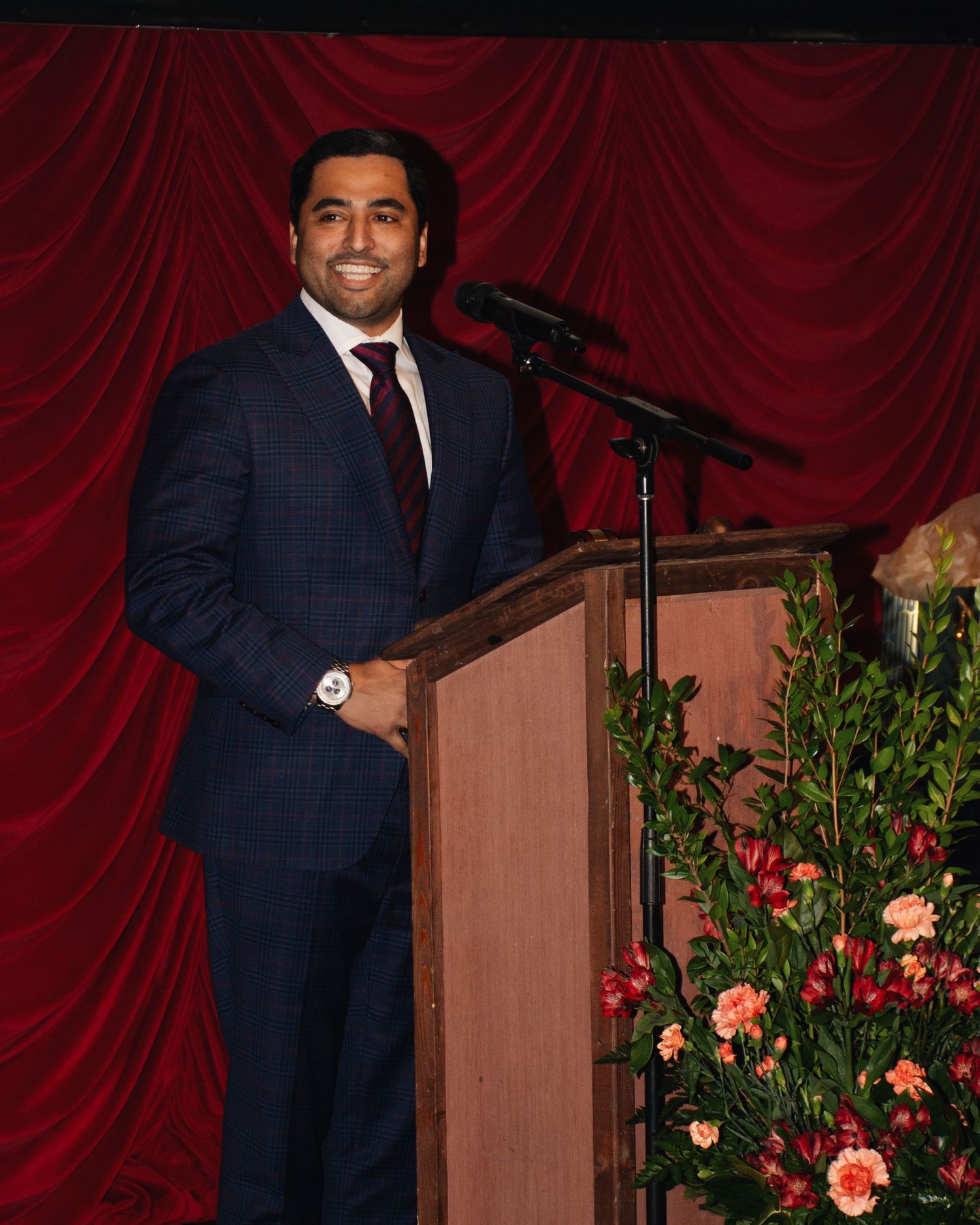 A speaker stands at a wooden podium with red curtains and floral arrangements in the background.