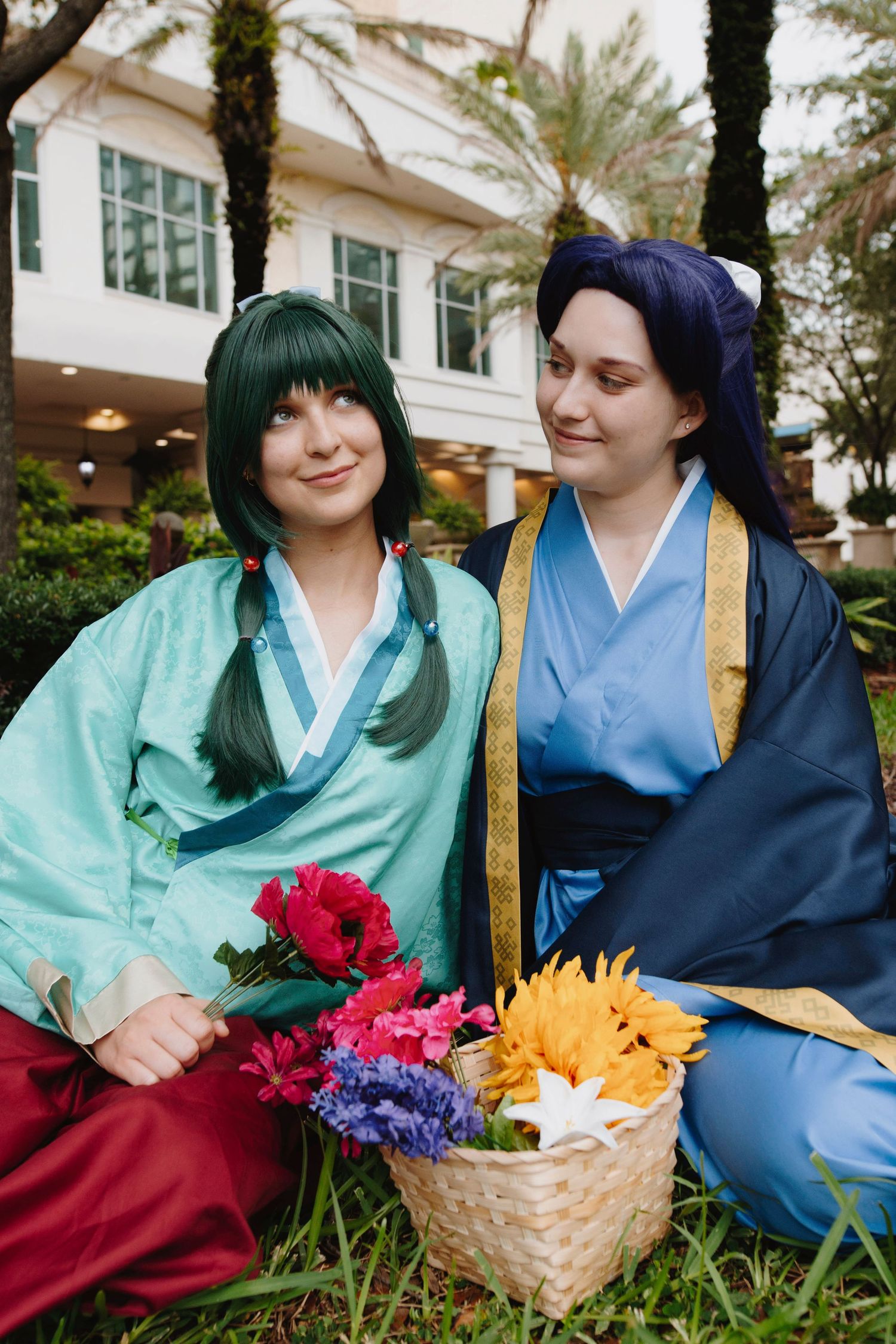 Two cosplayers in traditional Asian robes sit with flower basket in garden setting.
