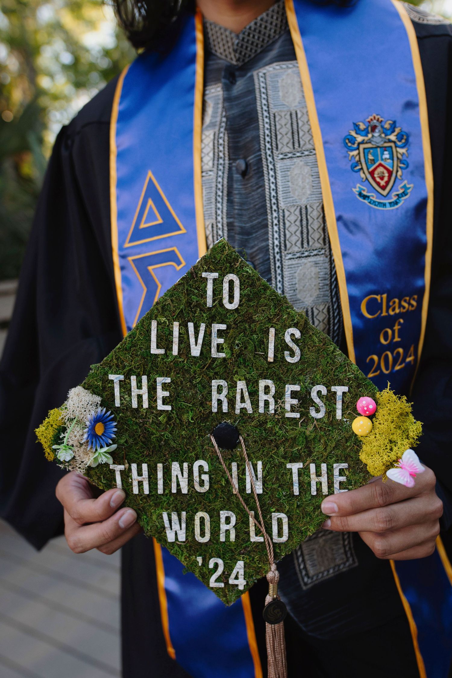Decorated graduation cap with moss lettering reads 'To live is the rarest thing in the world'.