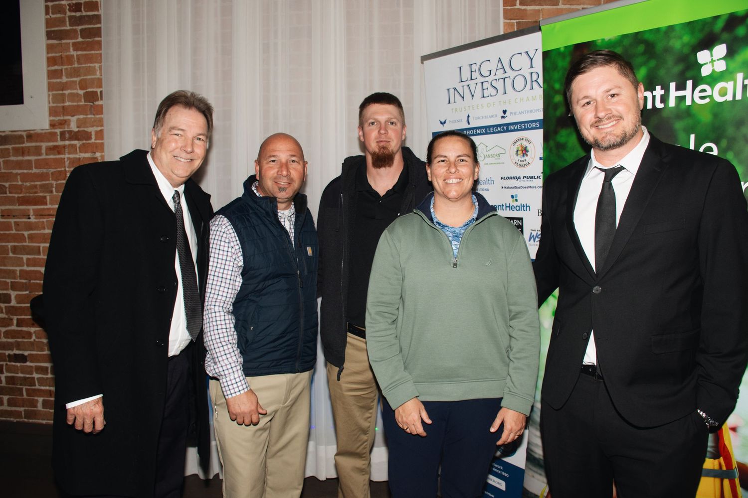 A group of people stand together in a line at a business event against a brick wall background.