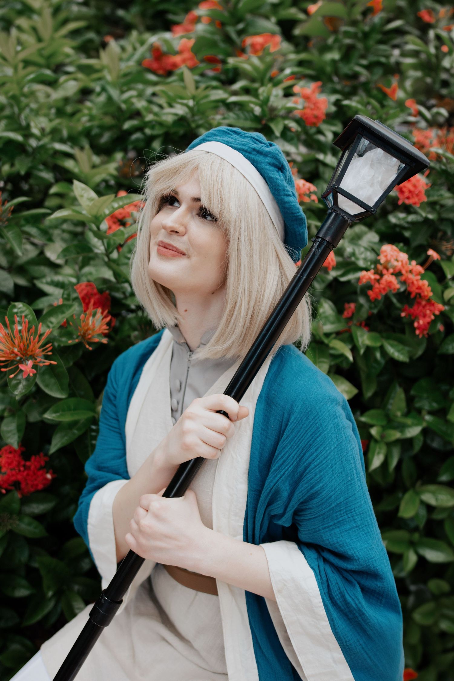 Cosplayer in blue cardigan poses playfully with prop sword against flowering red bush background.