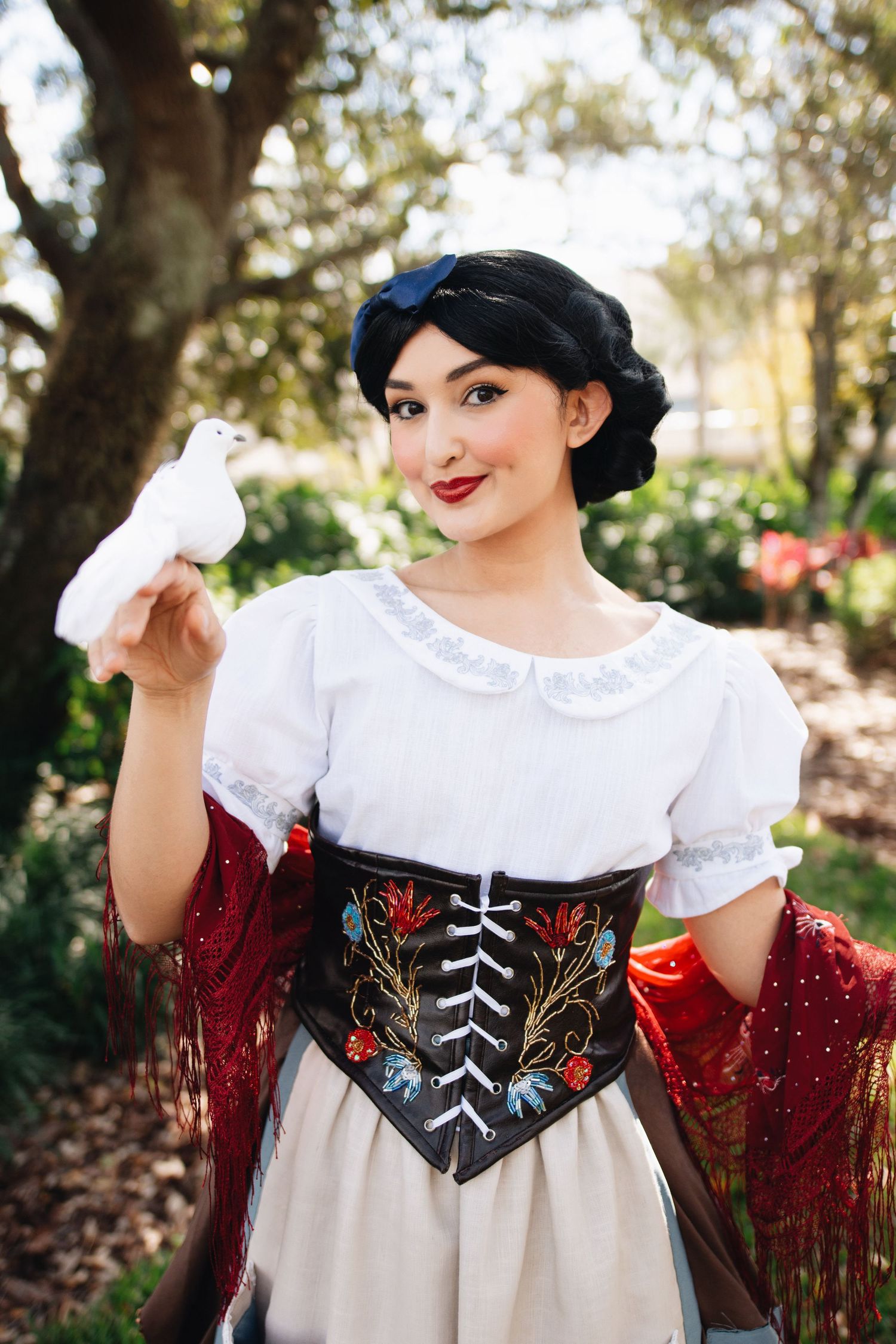 A historical costume featuring white blouse, embroidered corset and decorative red shawl.