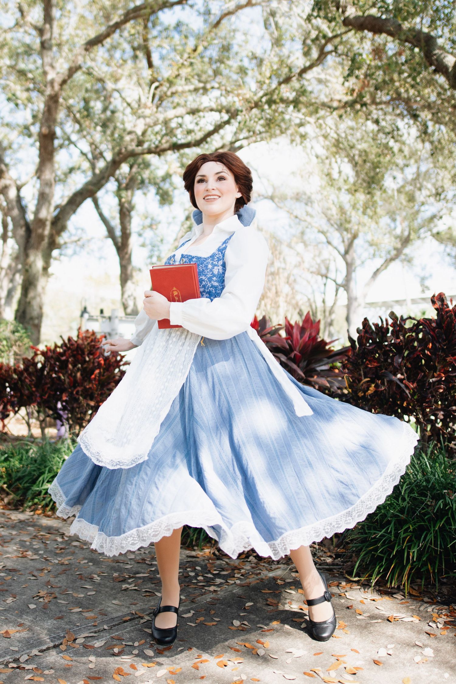 A cosplayer in a blue and white vintage dress twirls gracefully in a garden setting with trees in the background.