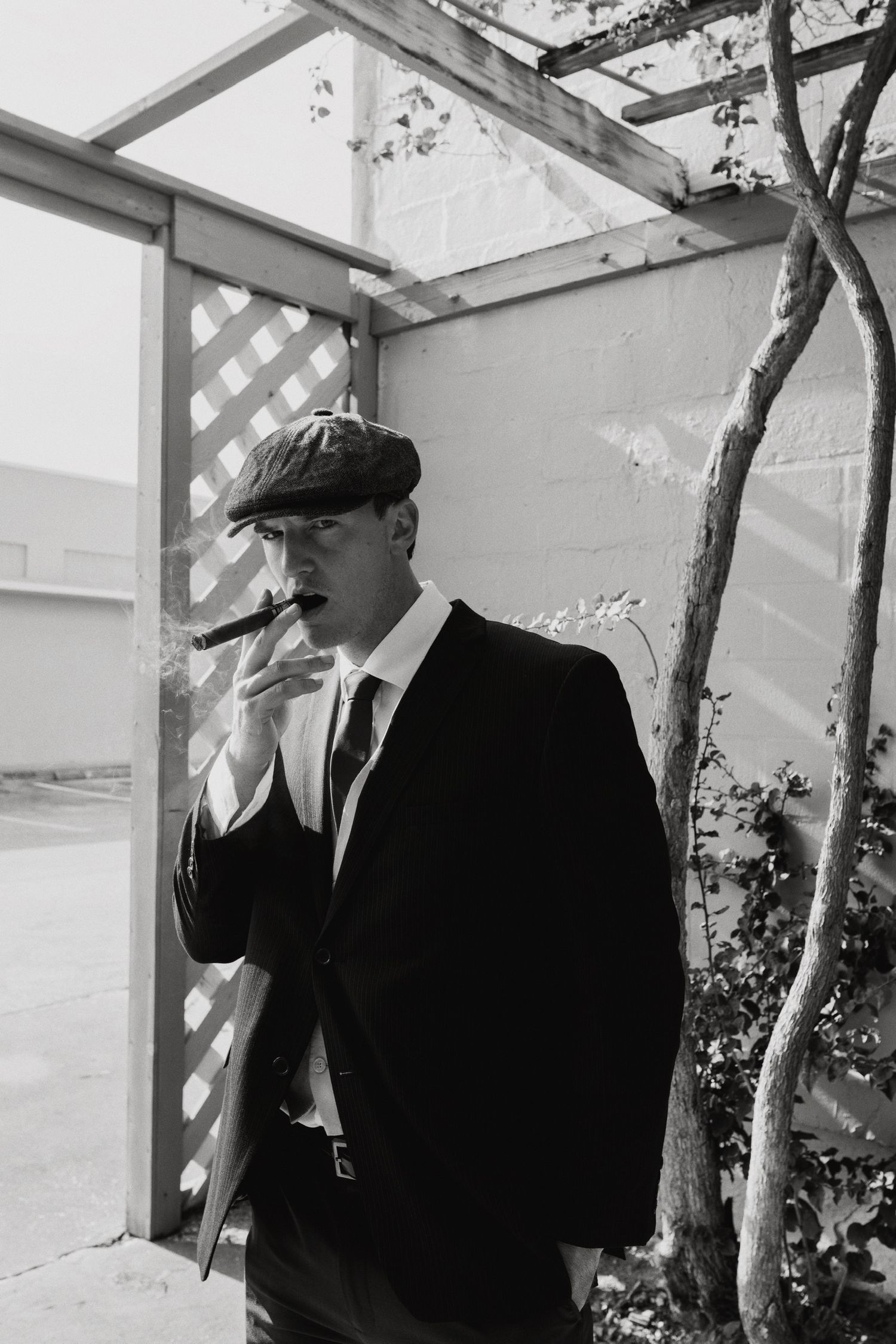 Vintage-style black and white portrait of person in suit and cap by latticed window.