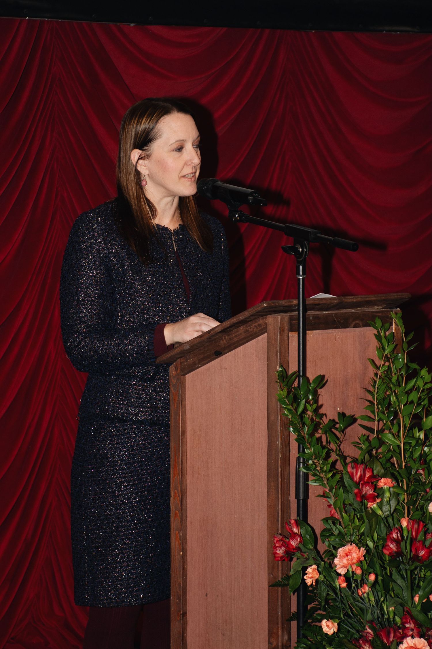 A speaker in a dark sparkly dress delivers remarks at a wooden podium with floral decorations against red curtains.