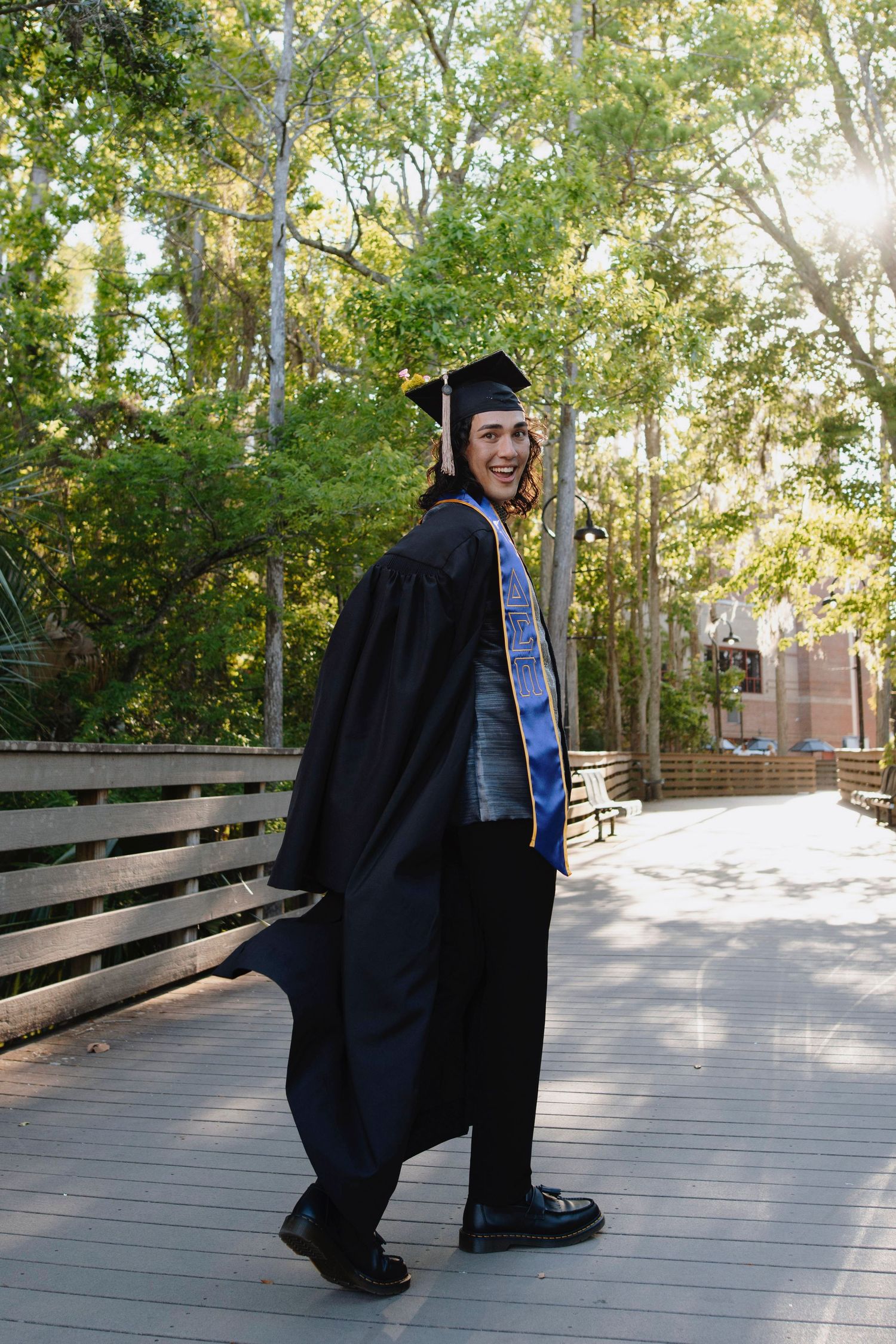 A graduate in academic regalia poses playfully on a wooden boardwalk surrounded by trees on a sunny day.
