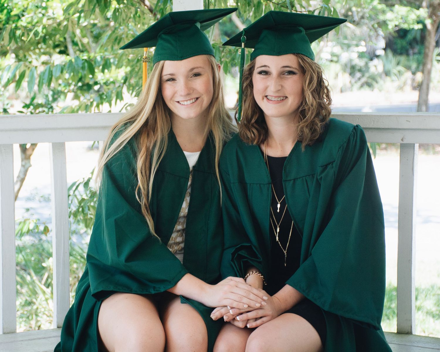 Two graduates in dark green caps and gowns sit together smiling on outdoor steps during commencement.