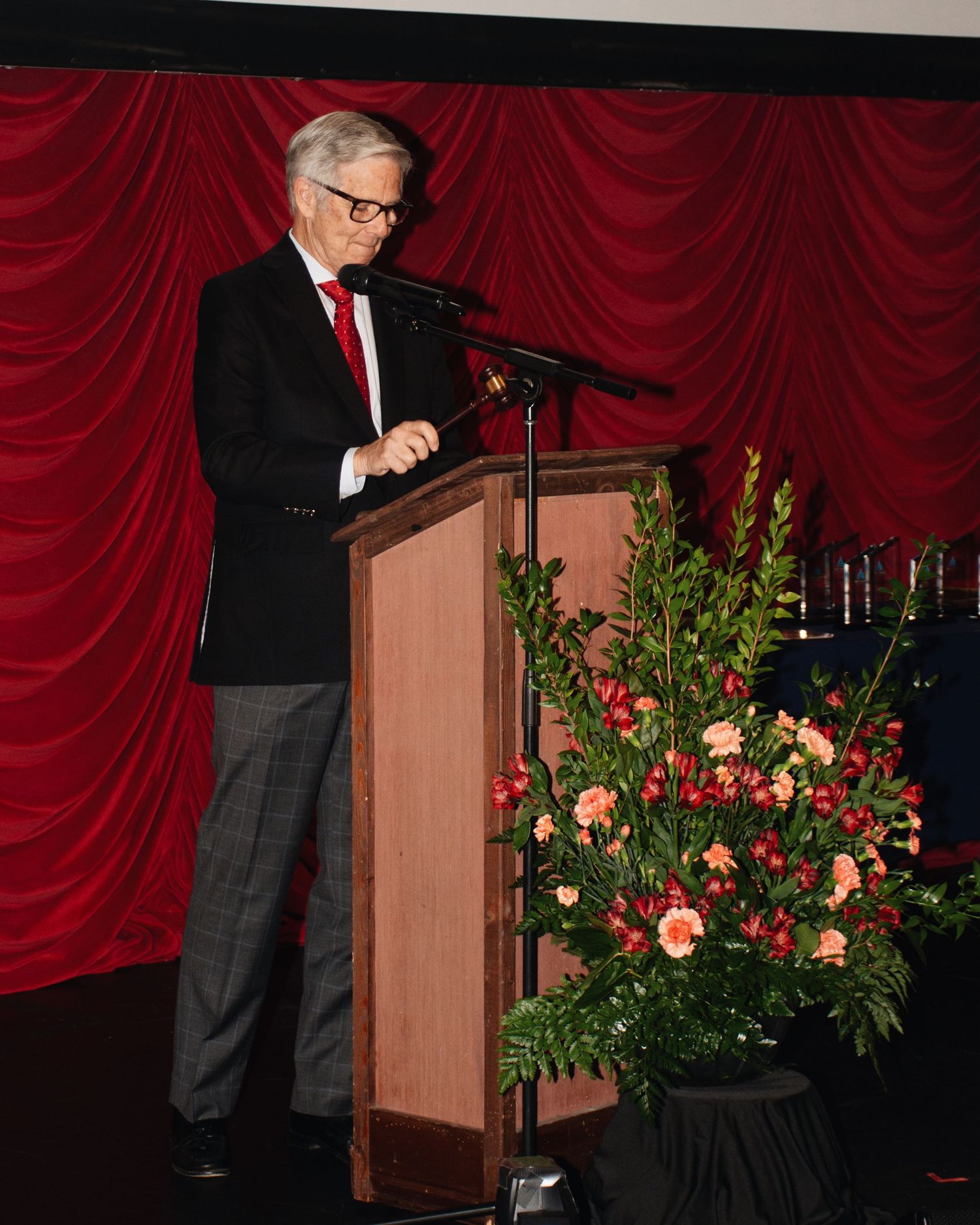 A speaker stands at a wooden podium with red curtains and floral arrangements in the background during a formal event.