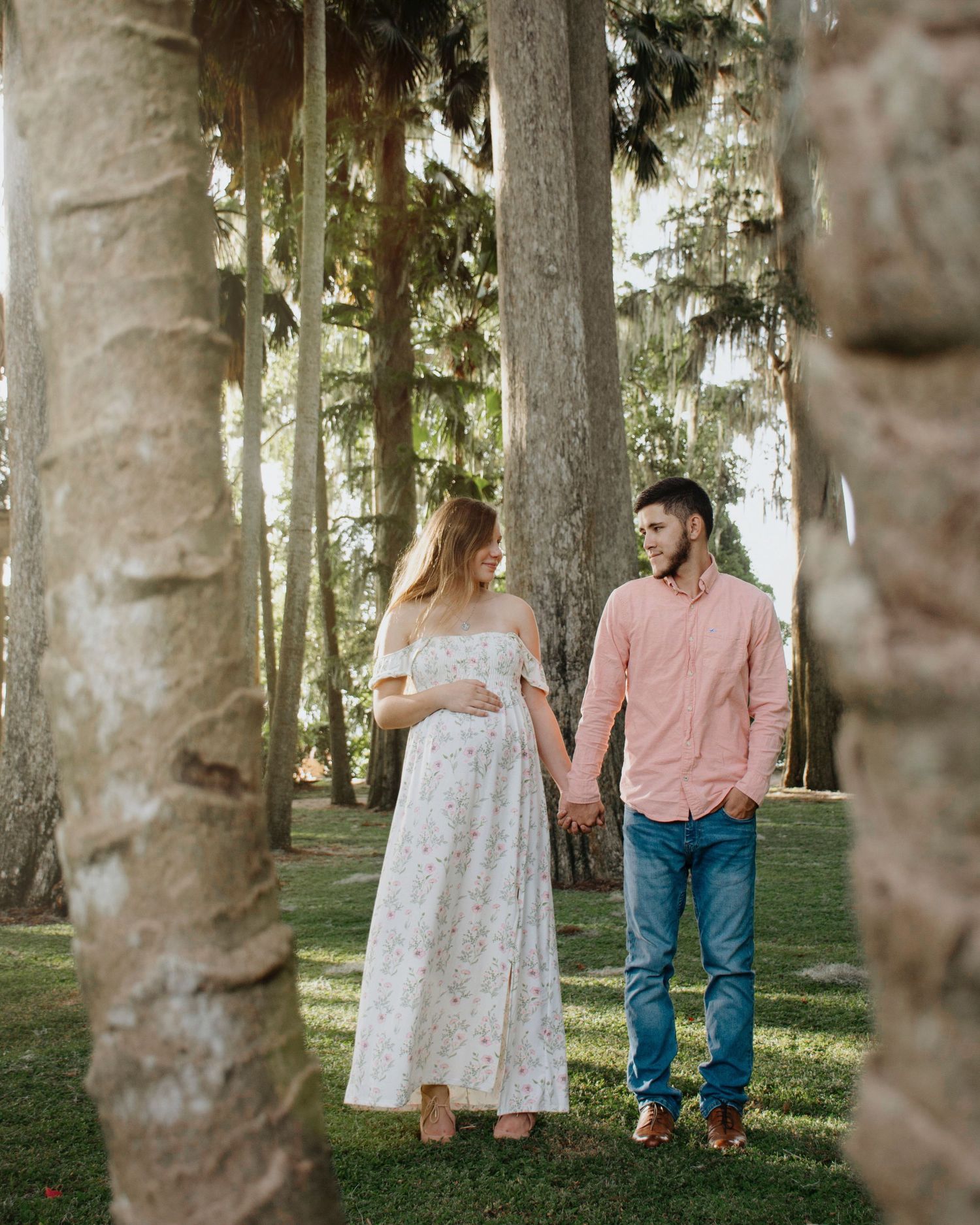 A couple walks hand in hand through a dreamy forest setting with tall trees and dappled sunlight.