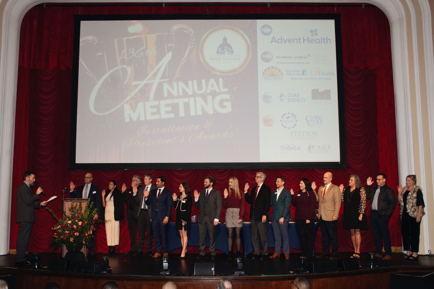 A large group of people in formal attire stand on stage during what appears to be an annual meeting or ceremony.