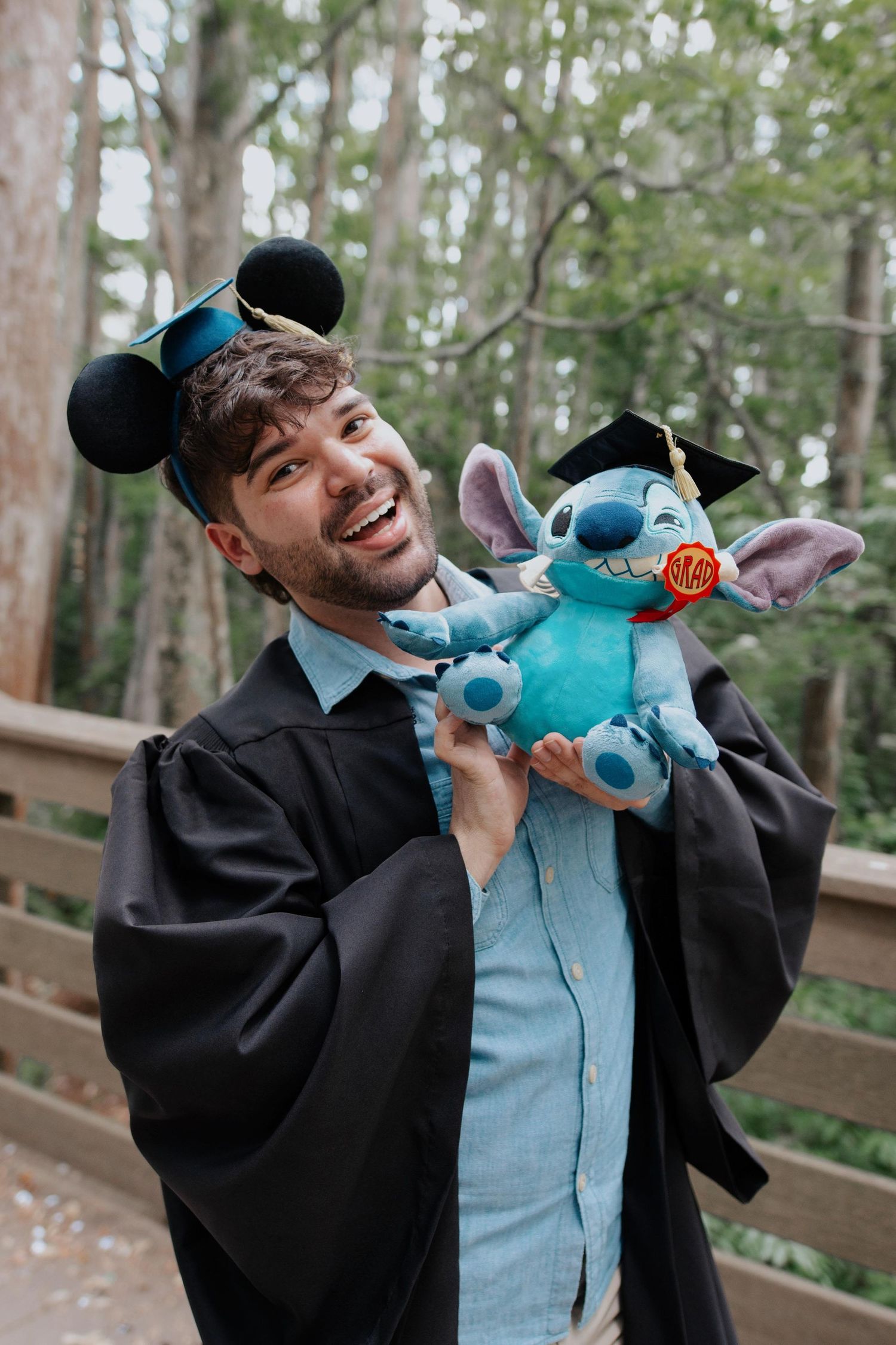 Graduate in cap and gown plays with blue plush toy in wooded setting.