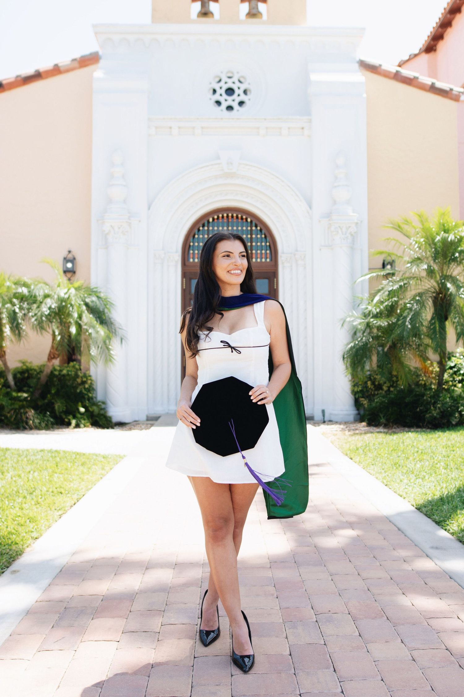 Graduate in white dress poses in front of Mediterranean-style building entrance.
