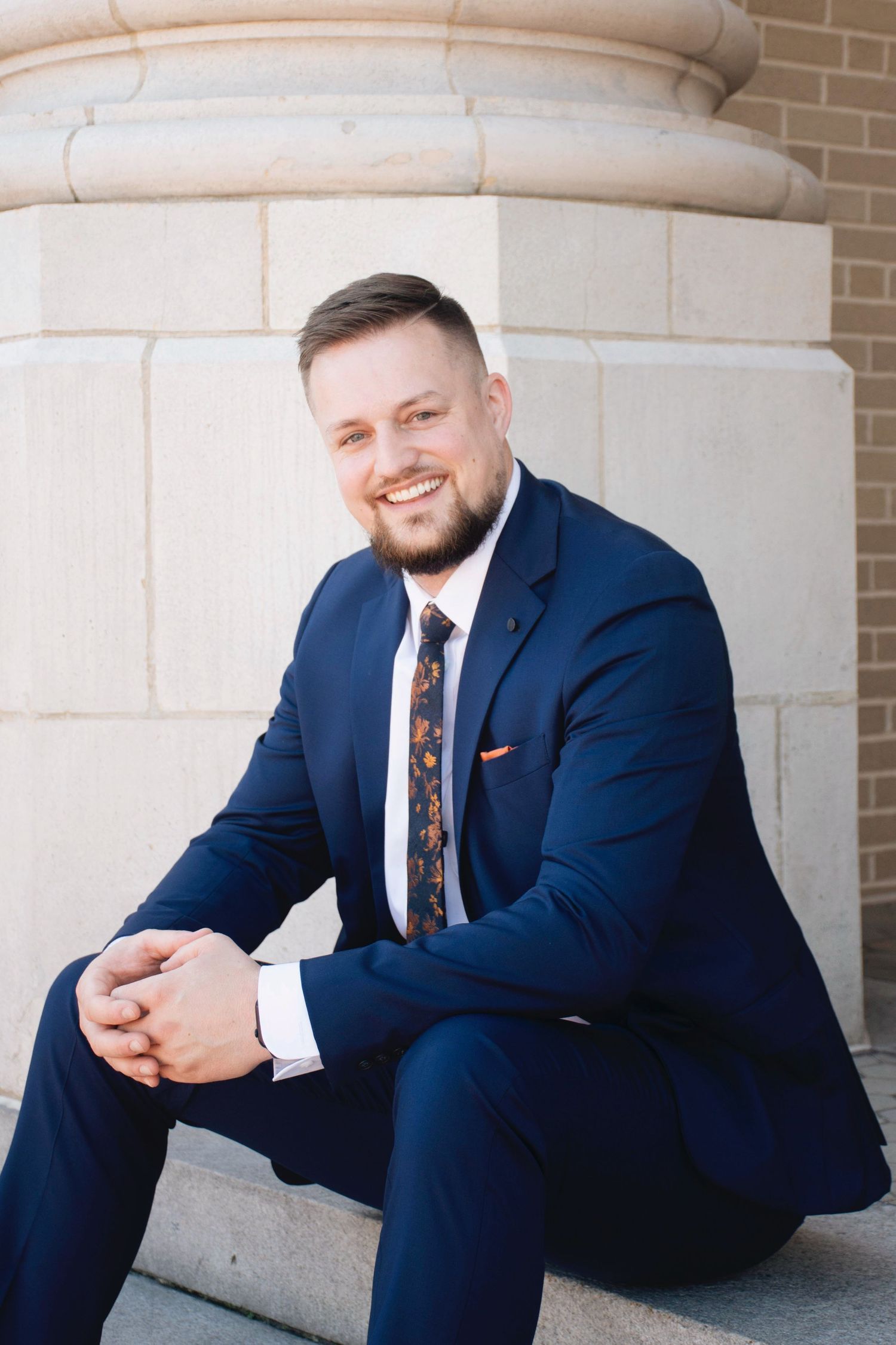 A person in a navy suit and tie poses casually against stone columns in a professional photoshoot.