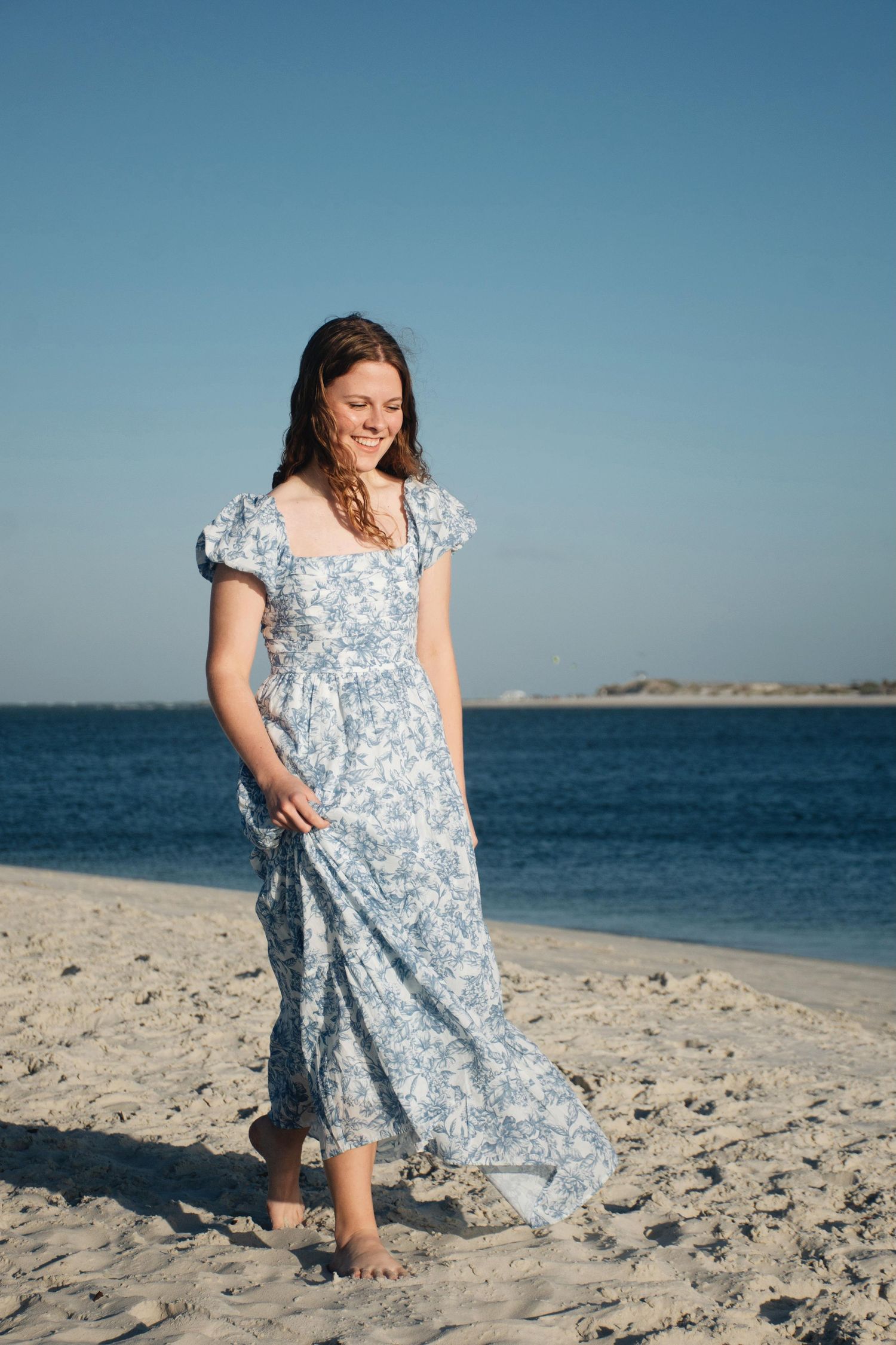Young woman in blue floral dress twirls on sandy beach with ocean backdrop.