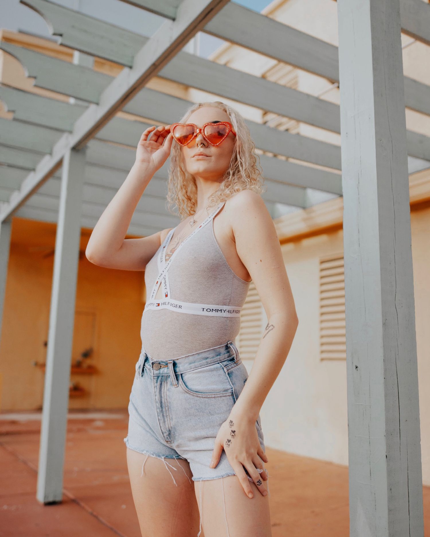 Fashion model poses in denim shorts and tank top against industrial backdrop with orange walls and beams.