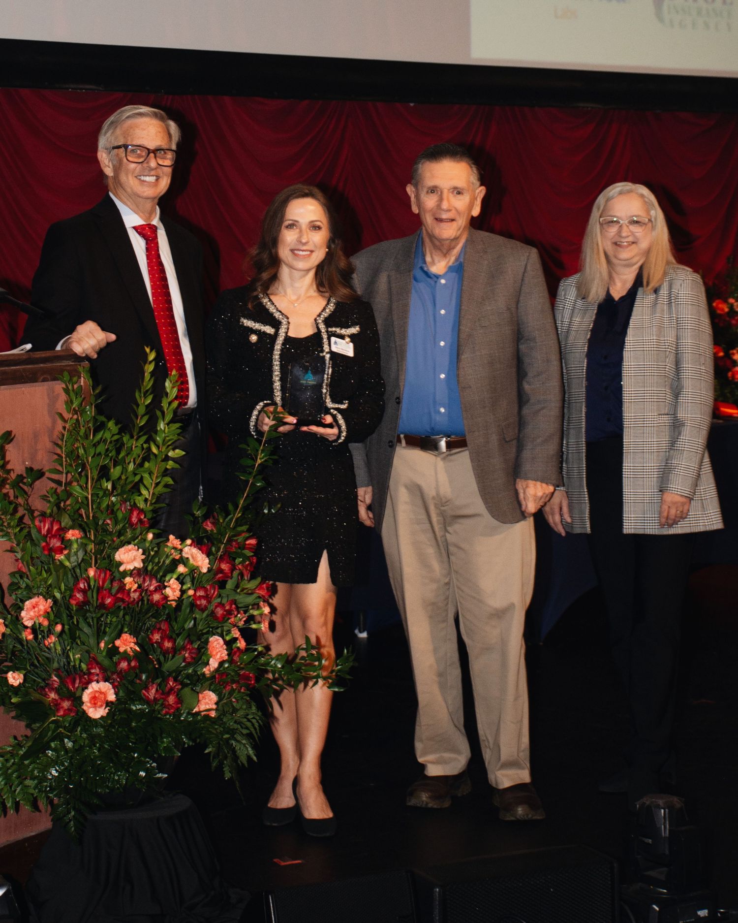 A group of people standing together at a formal event with red curtains and floral arrangements in the background.