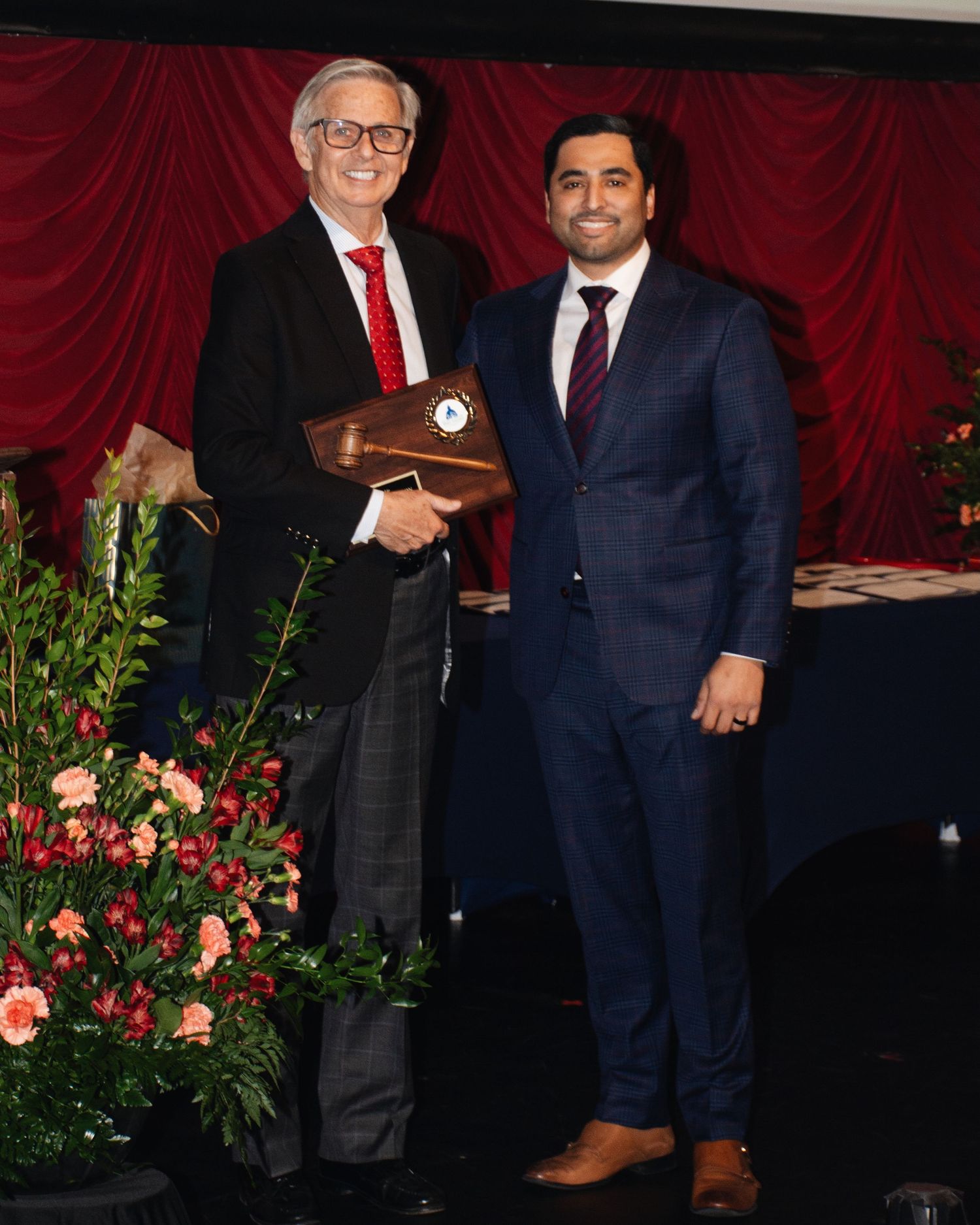 Two people in formal attire stand together at an awards ceremony with red curtains and floral decorations in the background.