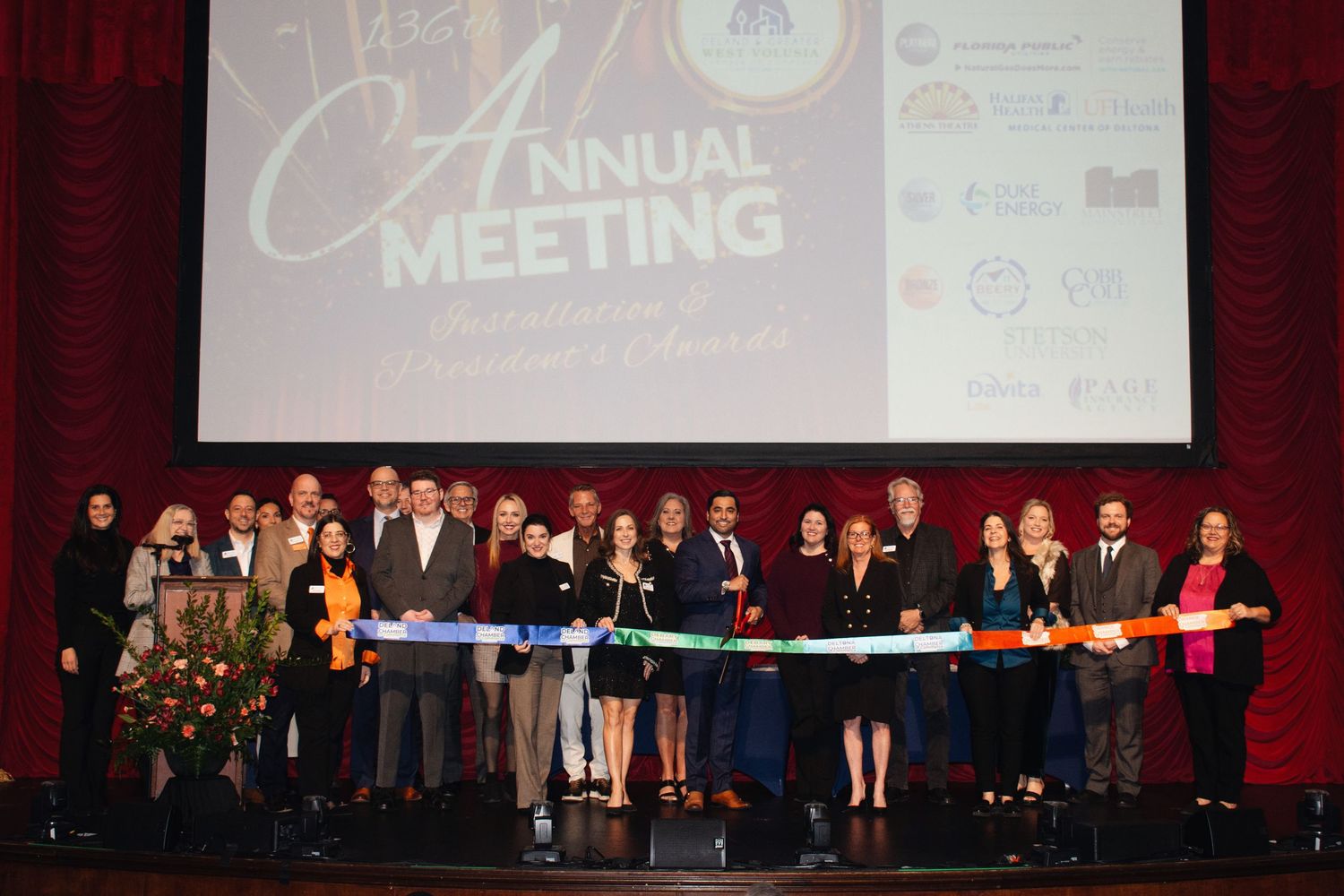 Conference attendees gather on stage during an annual meeting event with projection screens visible in background.