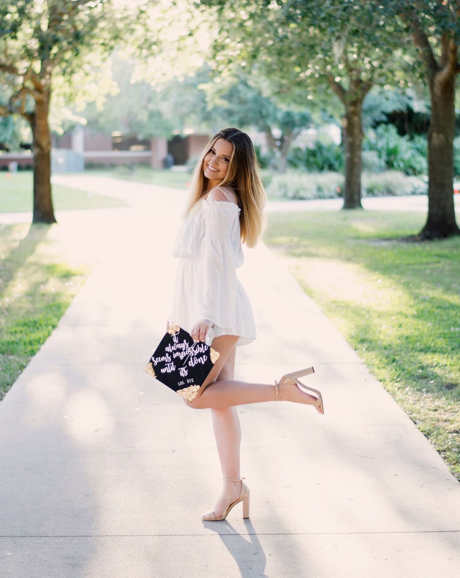 Graduate in white dress poses playfully on campus sidewalk holding decorated cap.