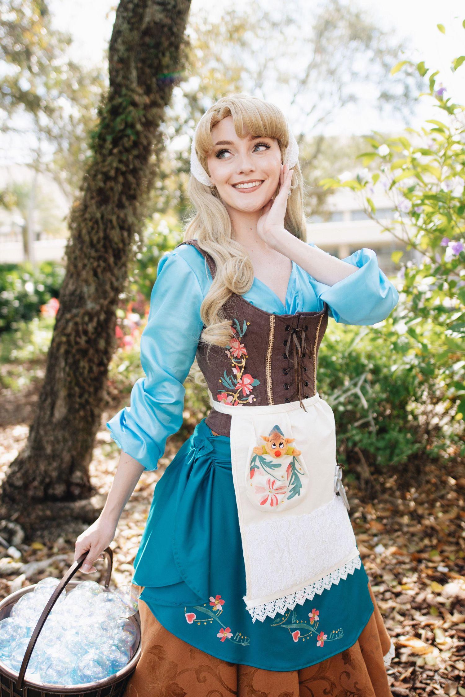 A costumed figure in a blue dress with brown corset and white apron poses with a basket in a garden.
