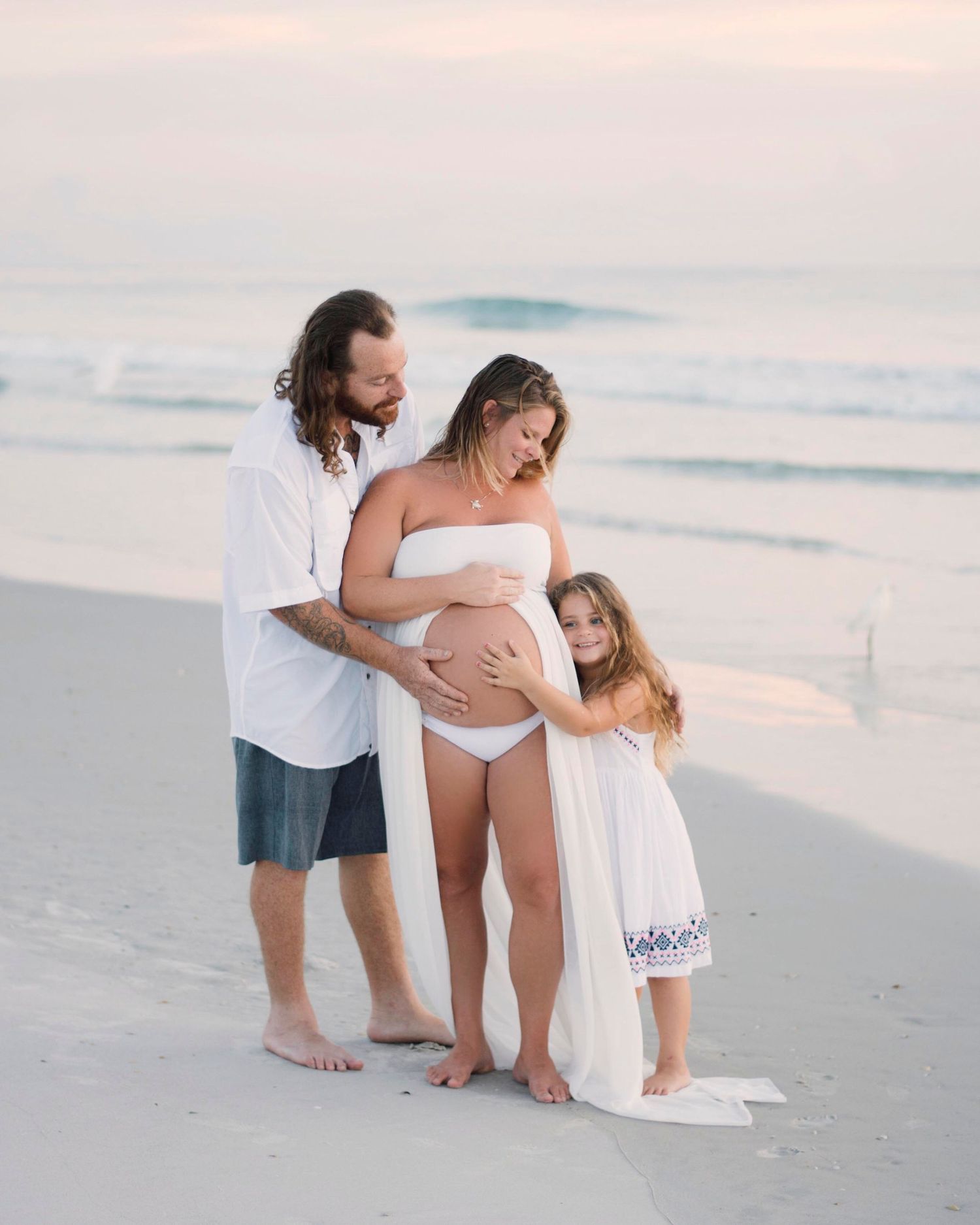 Family maternity photoshoot on a serene beach at sunset with white outfits and gentle poses.